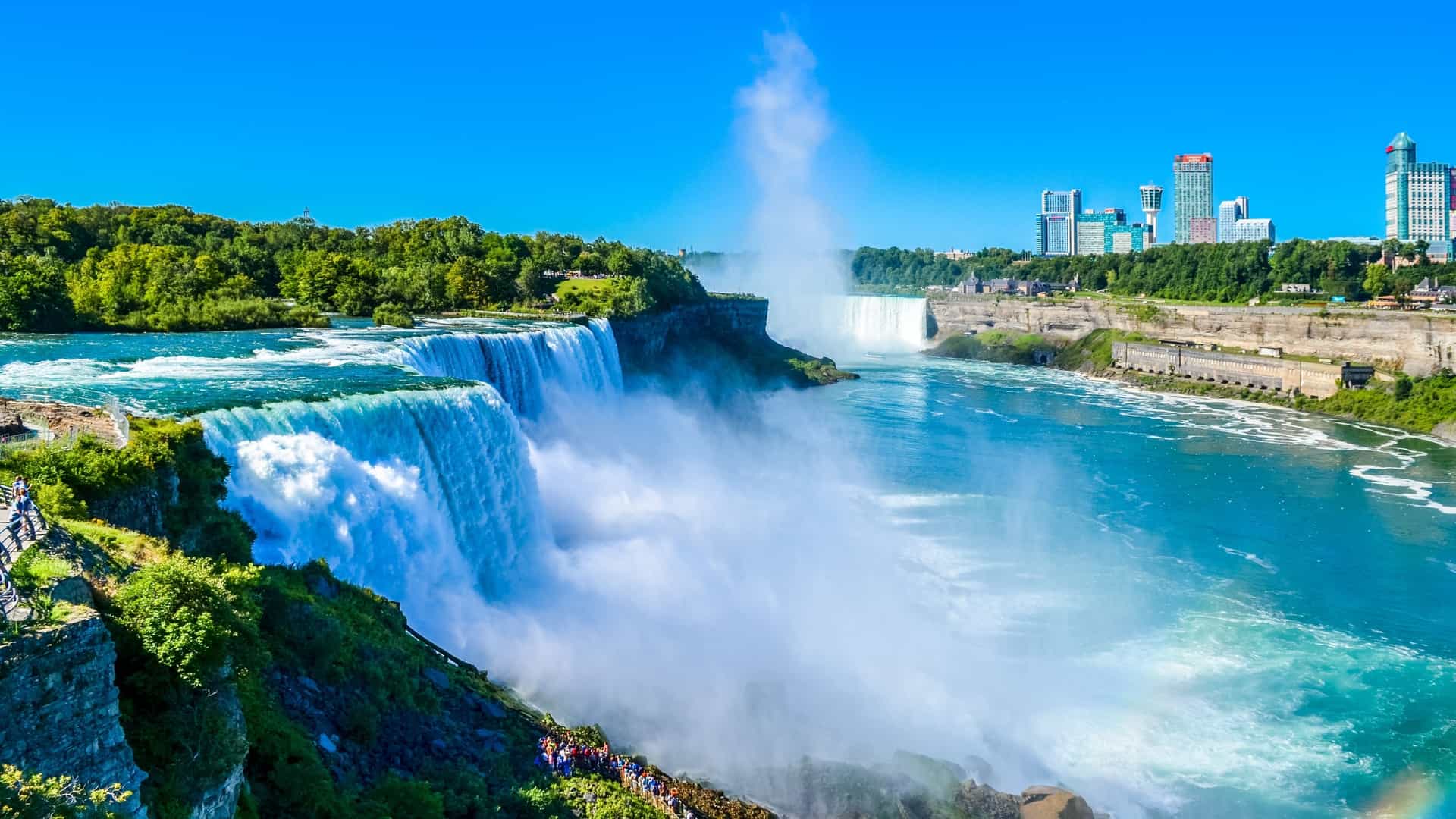 A spectacular high-angle view of the iconic Niagara Falls, a world-famous natural wonder and premier shore excursion destination situated near Niagara-on-the-Lake in Ontario, Canada. The image captures the immense volume of churning, turquoise-blue water of the American Falls plunging into a thick mist below, with the Horseshoe Falls visible in the background, all contrasted by lush green cliffs, a lower viewing pathway packed with visitors, and the modern city skyline of Niagara Falls rising against a clear blue sky.