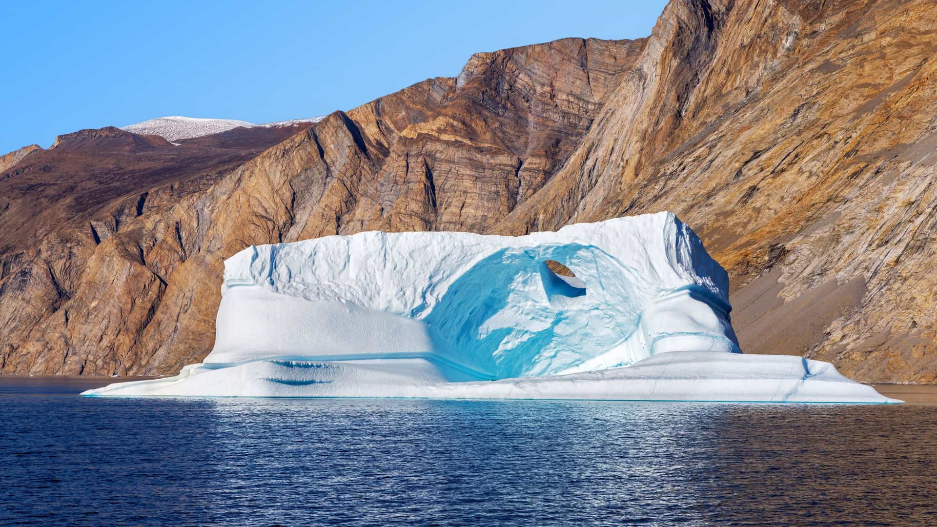 Large white iceberg with circular hollow floating in Arctic waters near rocky mountains under clear blue sky in Northeast Greenland National Park.