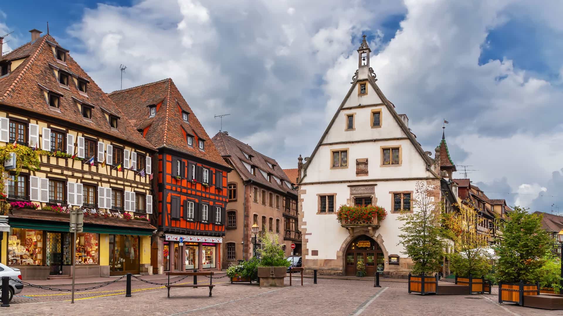 A colorful half‑timbered square in Obernai, France, with red‑and‑black and cream‑framed façades, flower‑lined windows, cobblestone streets, and planters under a bright sky, showcasing the medieval charm of this Alsace excursion stop.