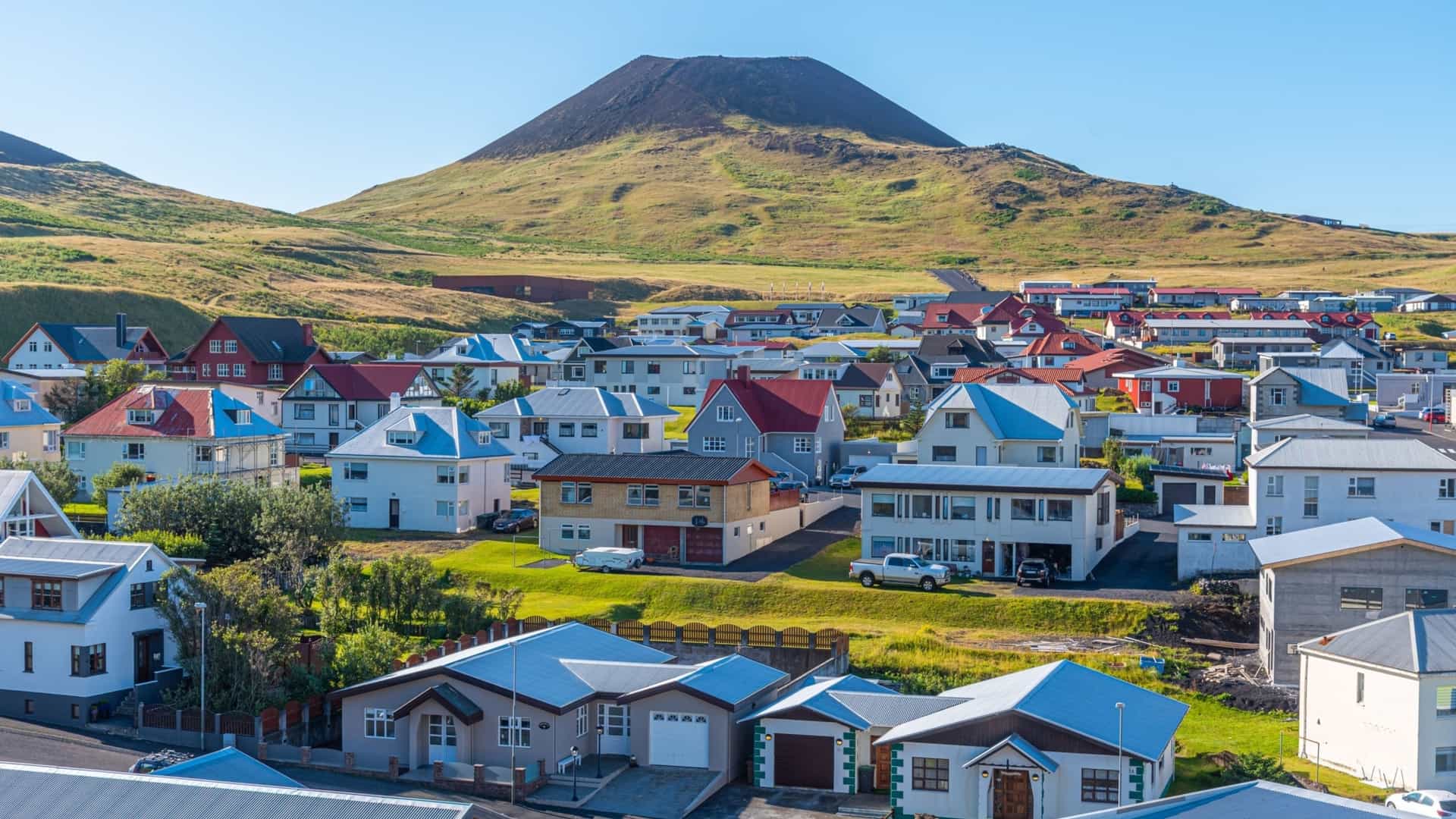 A picturesque eye-level view of a colorful residential neighborhood on the island of Heimaey, Iceland—a striking destination known for its dramatic volcanic history and maritime charm. The image features rows of well-maintained homes with varied roof colors—red, blue, and gray—set against a backdrop of a lush, grass-covered volcanic mountain cone under a clear, bright blue sky.