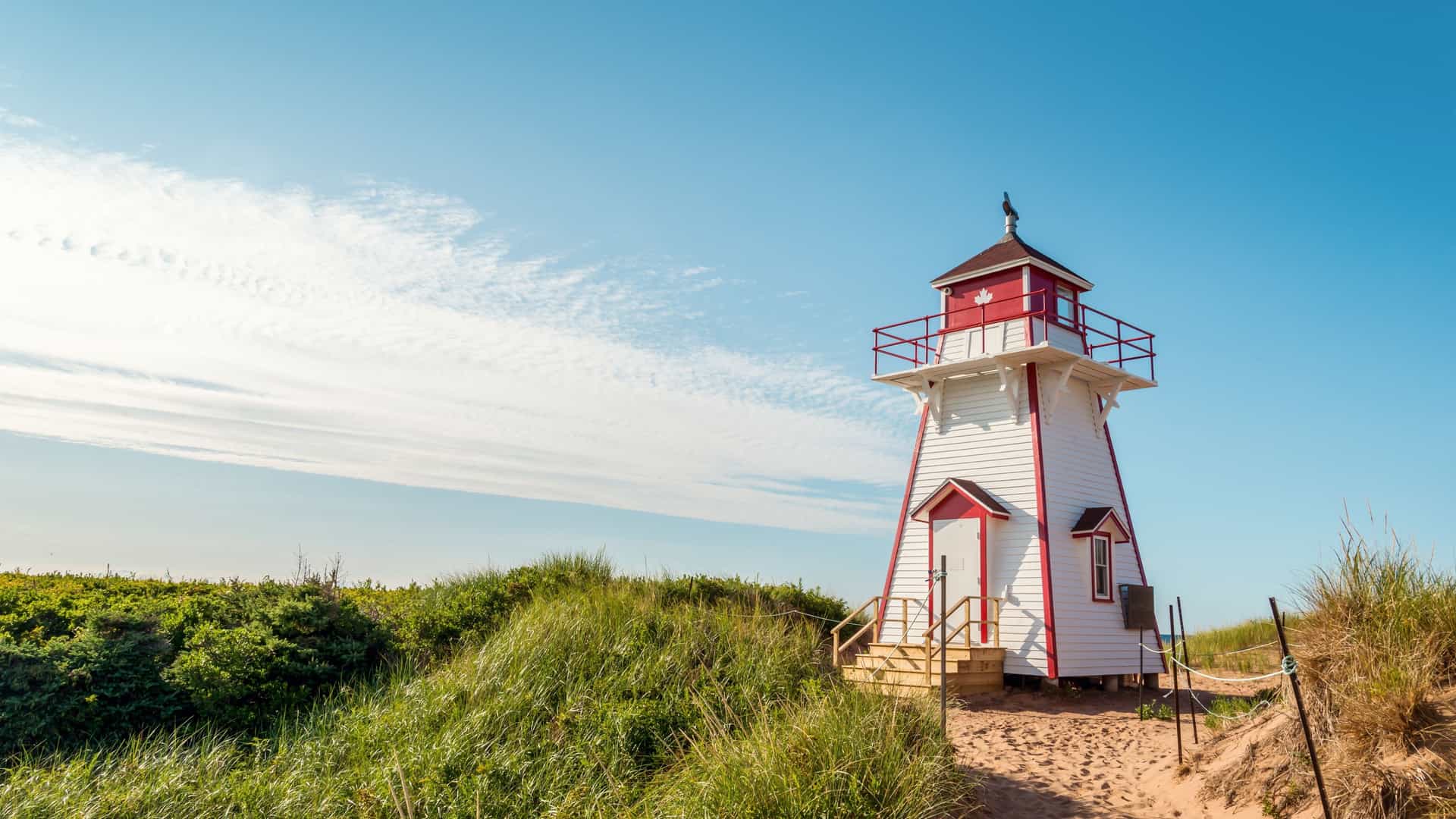 A red-and-white wooden lighthouse stands on a sandy path surrounded by green dune grass under a bright blue sky on Prince Edward Island, Canada.