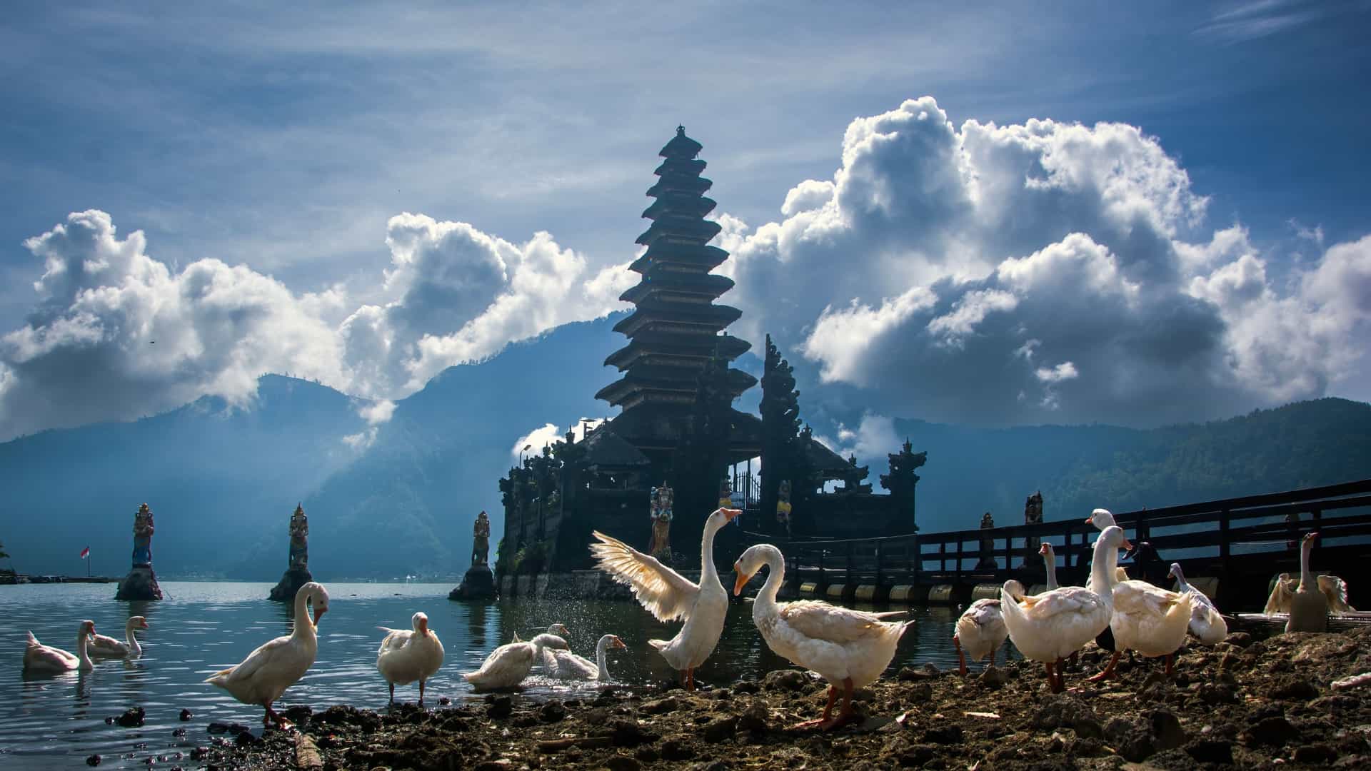 A lakeside Pulau Swande temple with multi‑tiered Balinese architecture, white ducks along a rocky shore, and misty forested mountains under dramatic clouds.