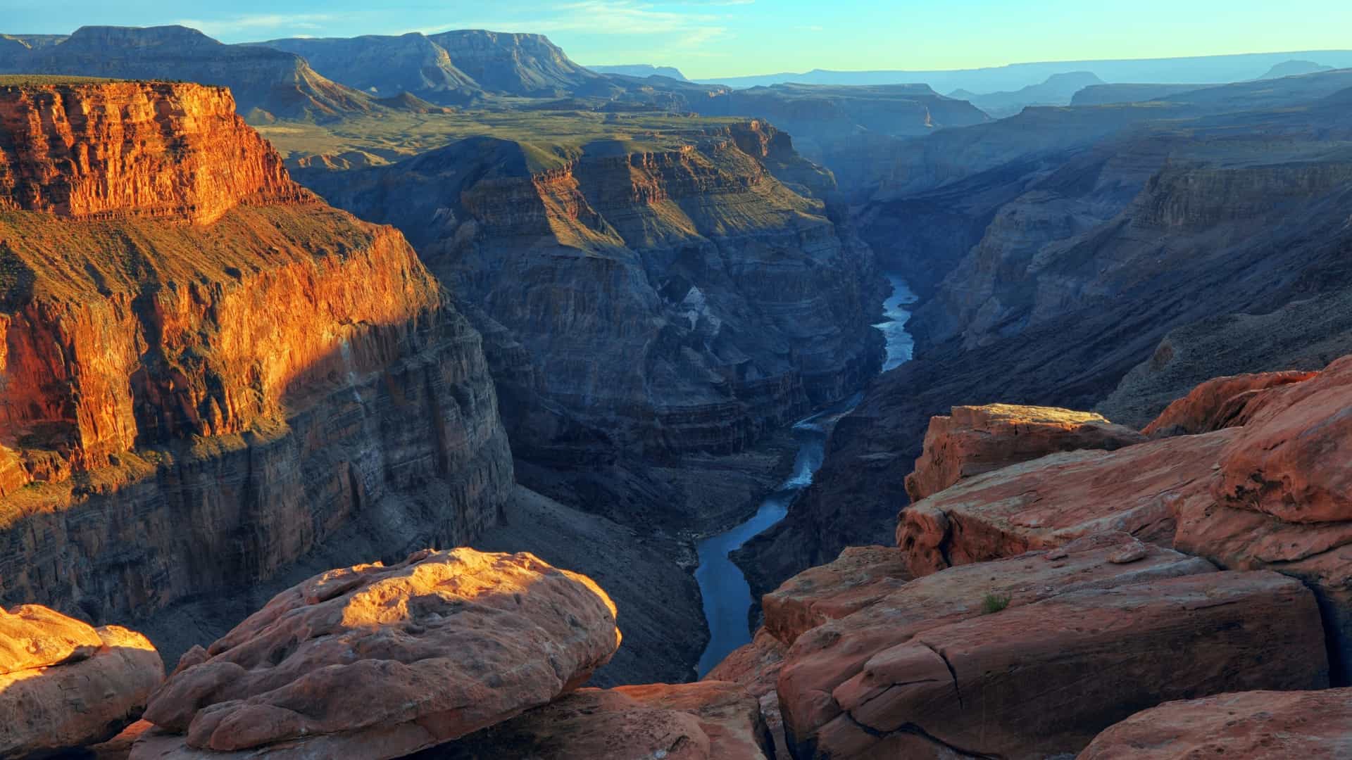 A deep canyon with a winding river is surrounded by towering, golden-lit red rock cliffs in Australia.