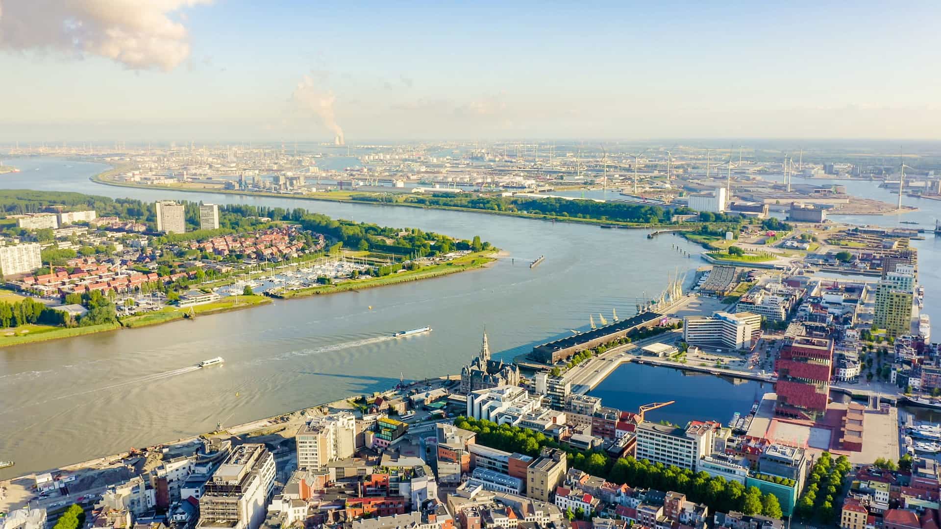 An aerial view of the Scheldt River with barges and boats moving along its curved waterway, dense urban districts on both banks, a marina and modern buildings in the foreground, and a vast industrial port with cranes and wind turbines stretching into the distance.