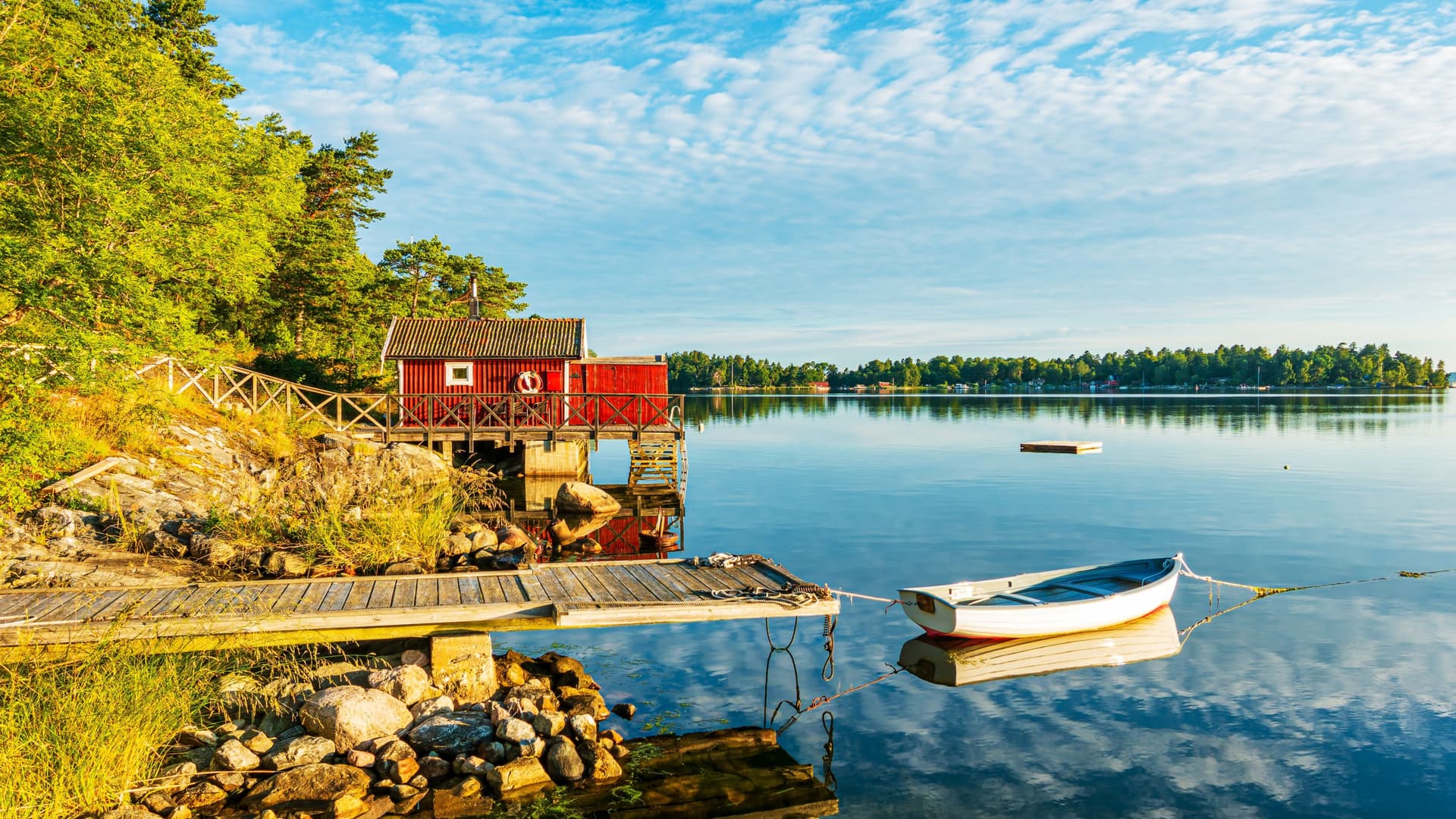 A red wooden cabin on stilts along the calm waters of the Stockholm Archipelago, with a small dock, a moored rowboat, and forested islands under a bright partly cloudy sky, capturing the serene coastal beauty of this Swedish island chain.