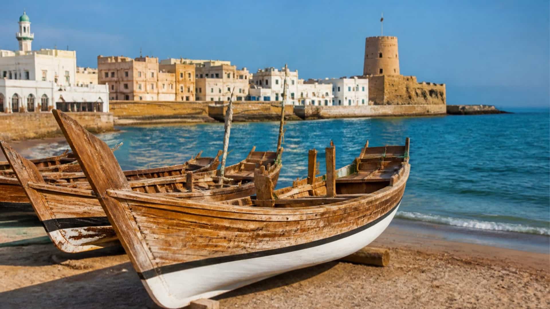 Traditional Omani dhow boats rest on the beach at Sur, with the historic waterfront and the Arabian Sea beyond.