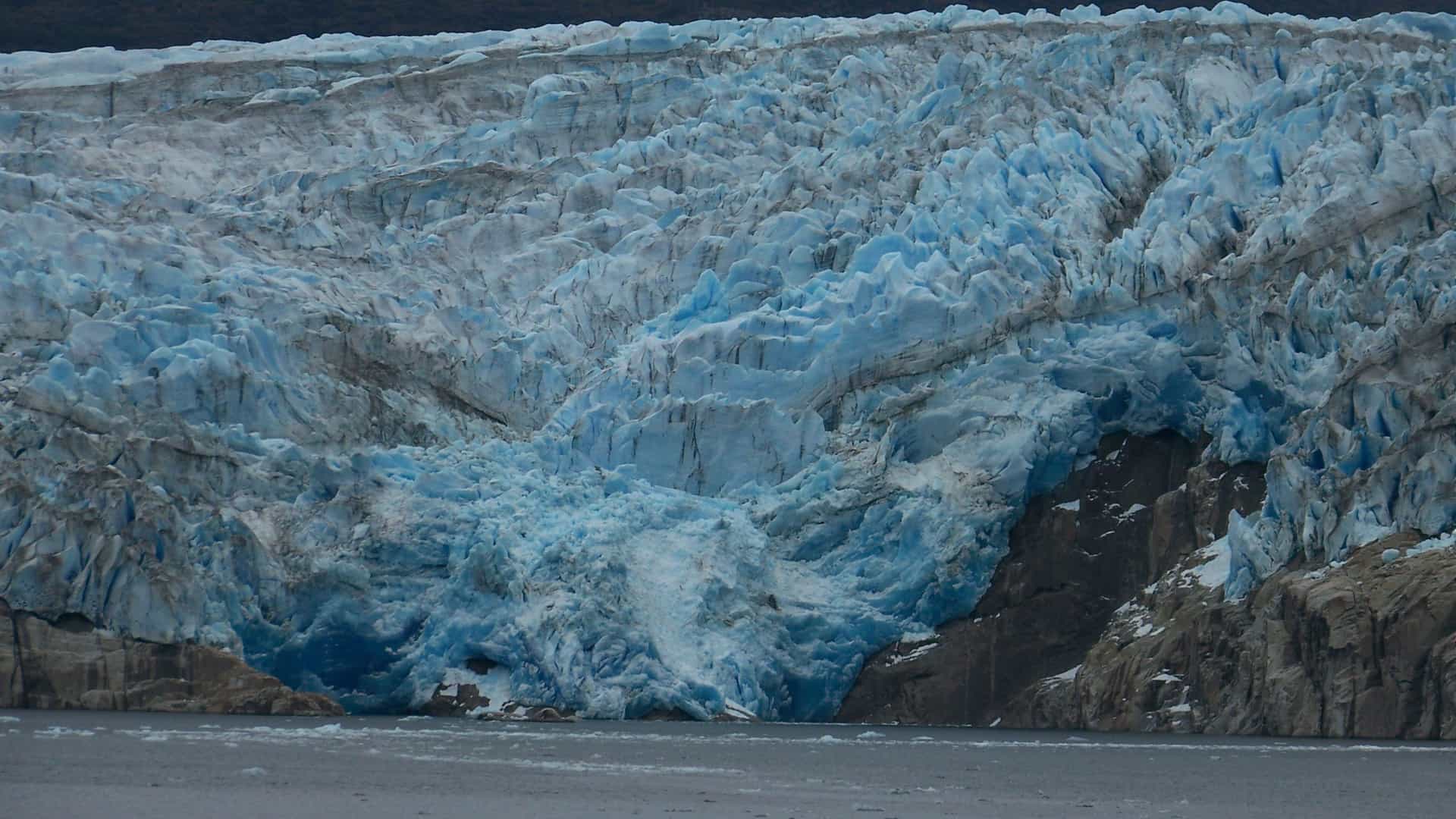Deep‑blue ice walls of the Tempanos Glacier with jagged crevasses and dark rocky outcrops descending into cold glacial water in Tierra del Fuego.