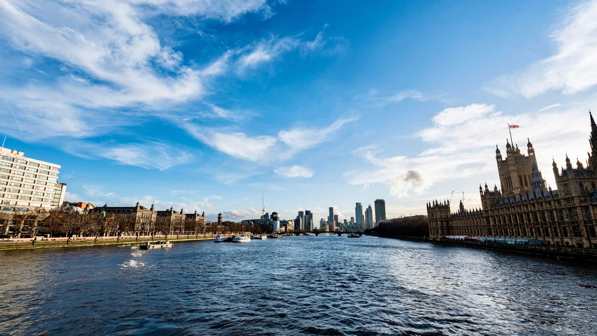 The River Thames with the Palace of Westminster on its bank, boats moving along the water, and London’s skyline stretching beneath a bright, partly cloudy sky.