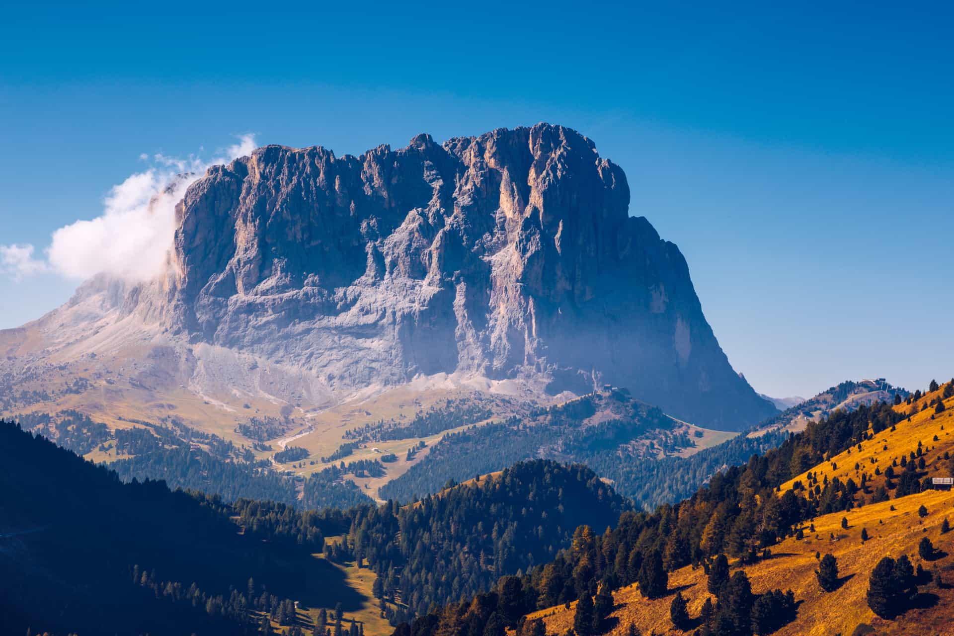 A massive, rugged mountain peak in the Dolomites rises above a forest-covered hillside under a clear blue sky.