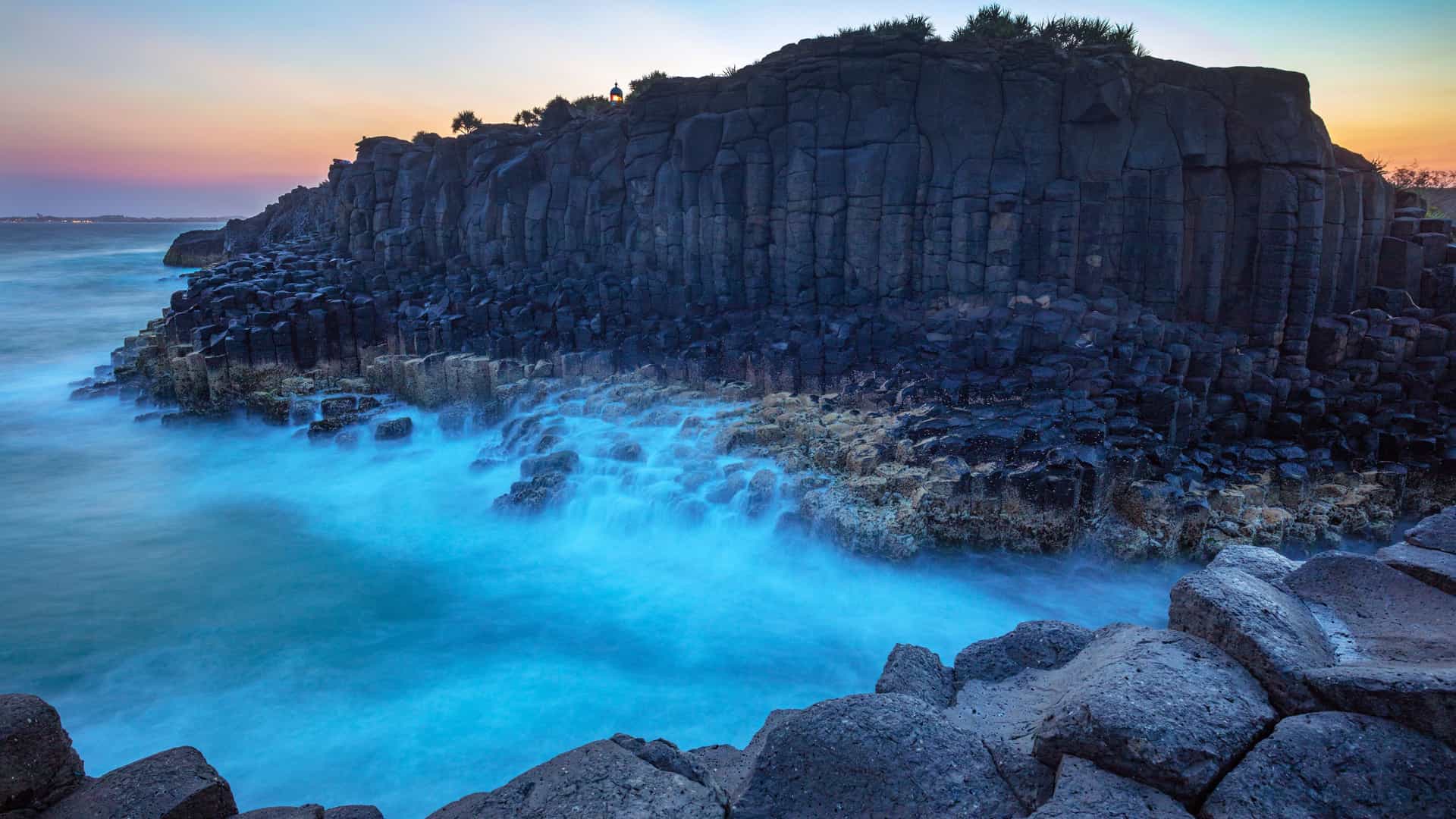 Tall hexagonal basalt columns rising along a rugged Australian coastline, with waves washing against the rock formations and a soft sunrise glow coloring the sky above the Far North Region.
