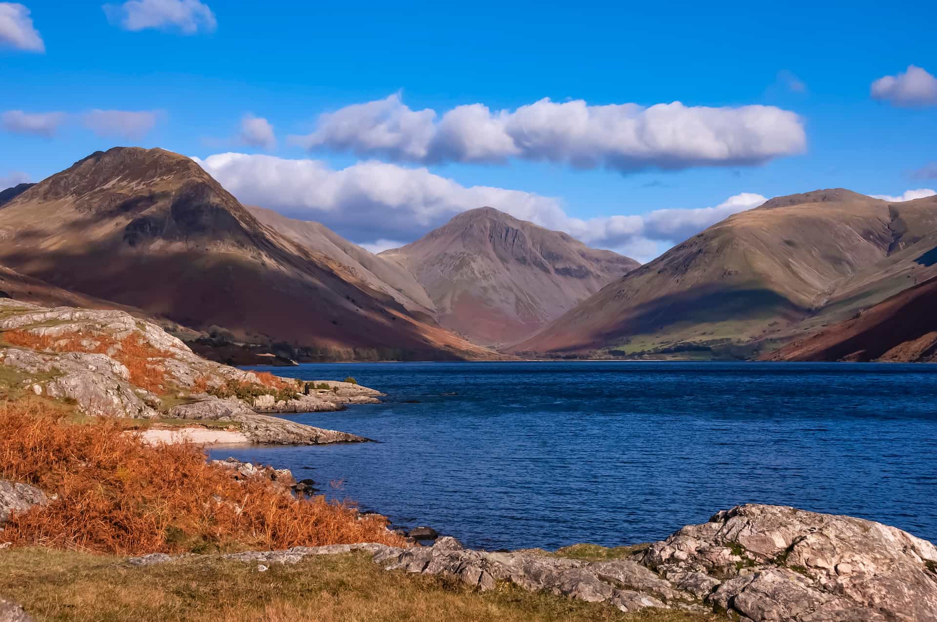 A deep blue lake surrounded by rolling, shadowed mountains under a bright, cloud-spotted sky.