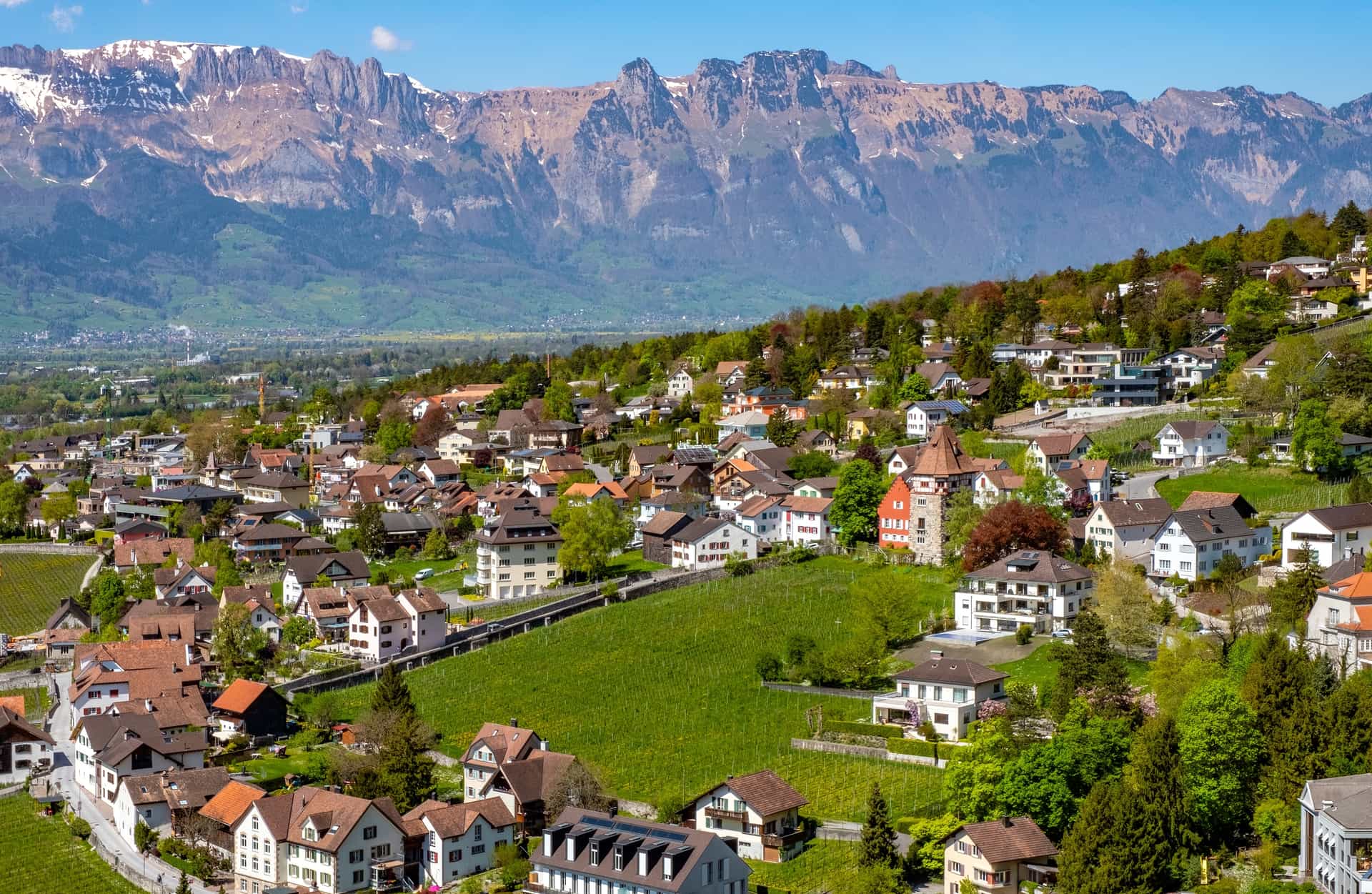 A scenic town nestled among lush green slopes, with a historic tower and expansive mountain ranges in the background.