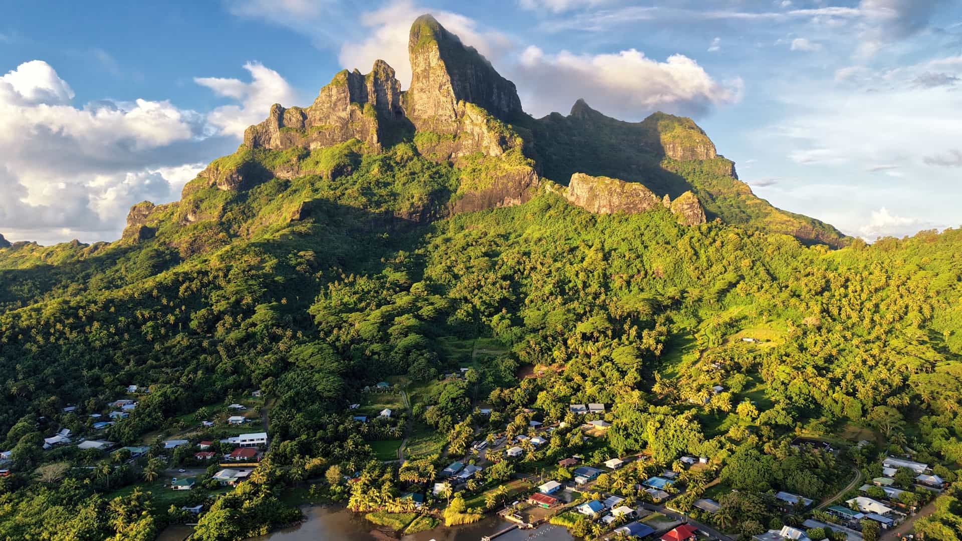 A dramatic view of Vaitape, Bora Bora, with steep, jagged green peaks rising above a small tropical settlement, sunlight highlighting the rugged volcanic landscape that defines this iconic French Polynesian island.