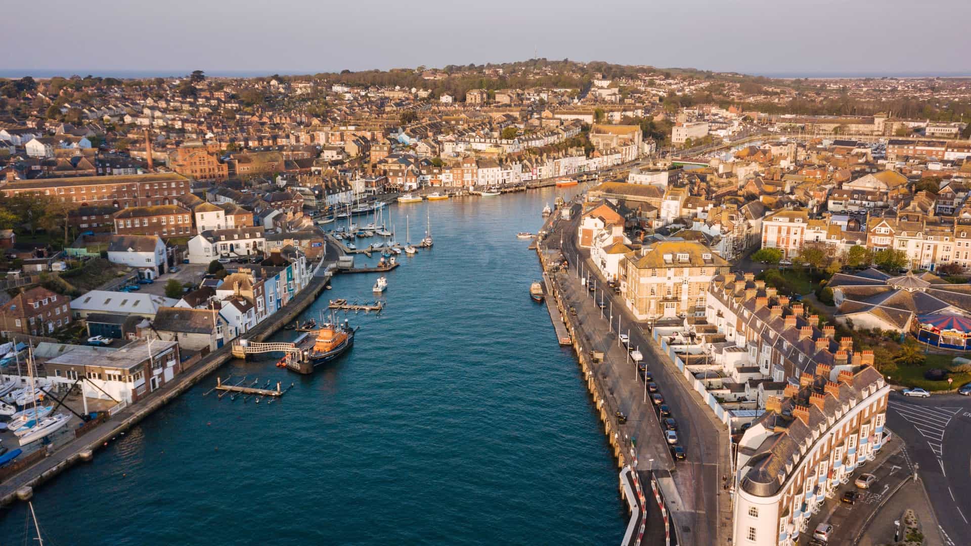 A warm aerial view of Weymouth, showing its central harbor lined with historic brick buildings, boats moored along the quays, and tightly packed homes stretching inland under late‑afternoon light.