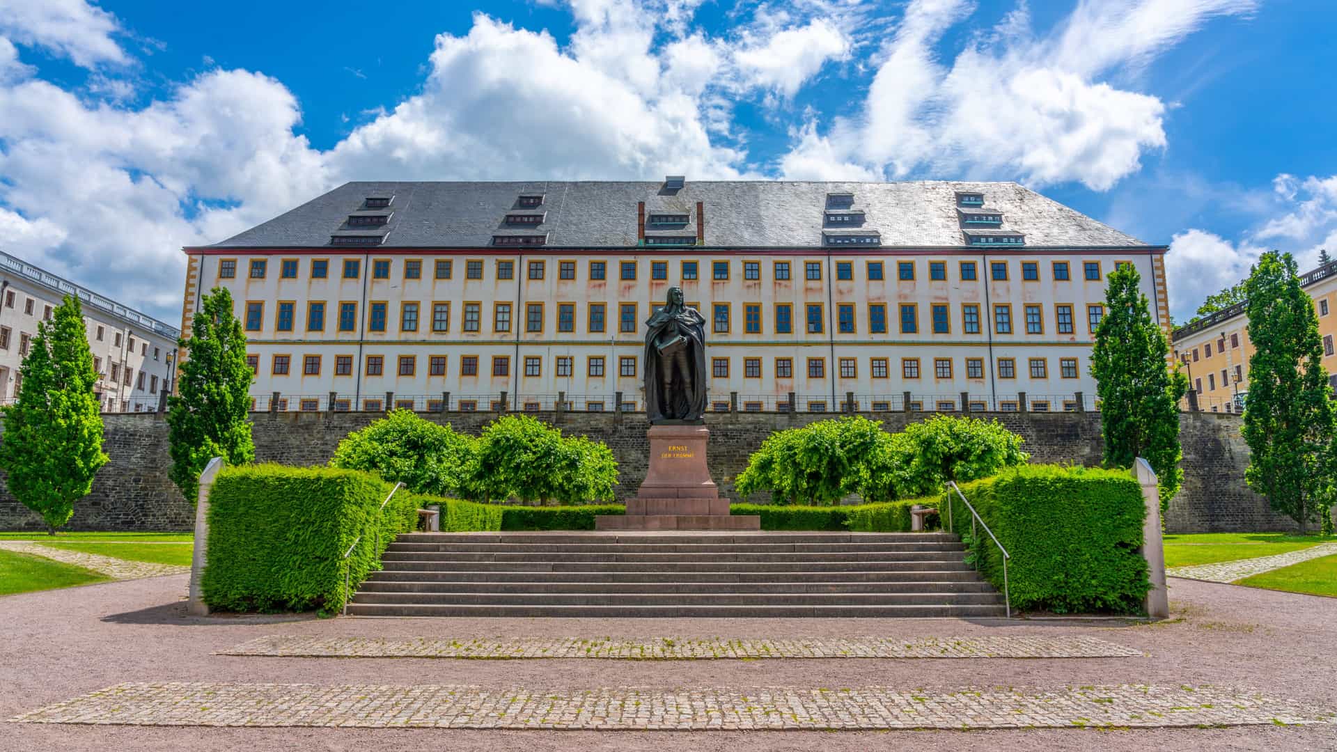 A historic building in Zons with a dark slate roof and rows of windows, fronted by the Ernst der Fromme statue, trimmed hedges, stone steps, and a partly cloudy blue sky.