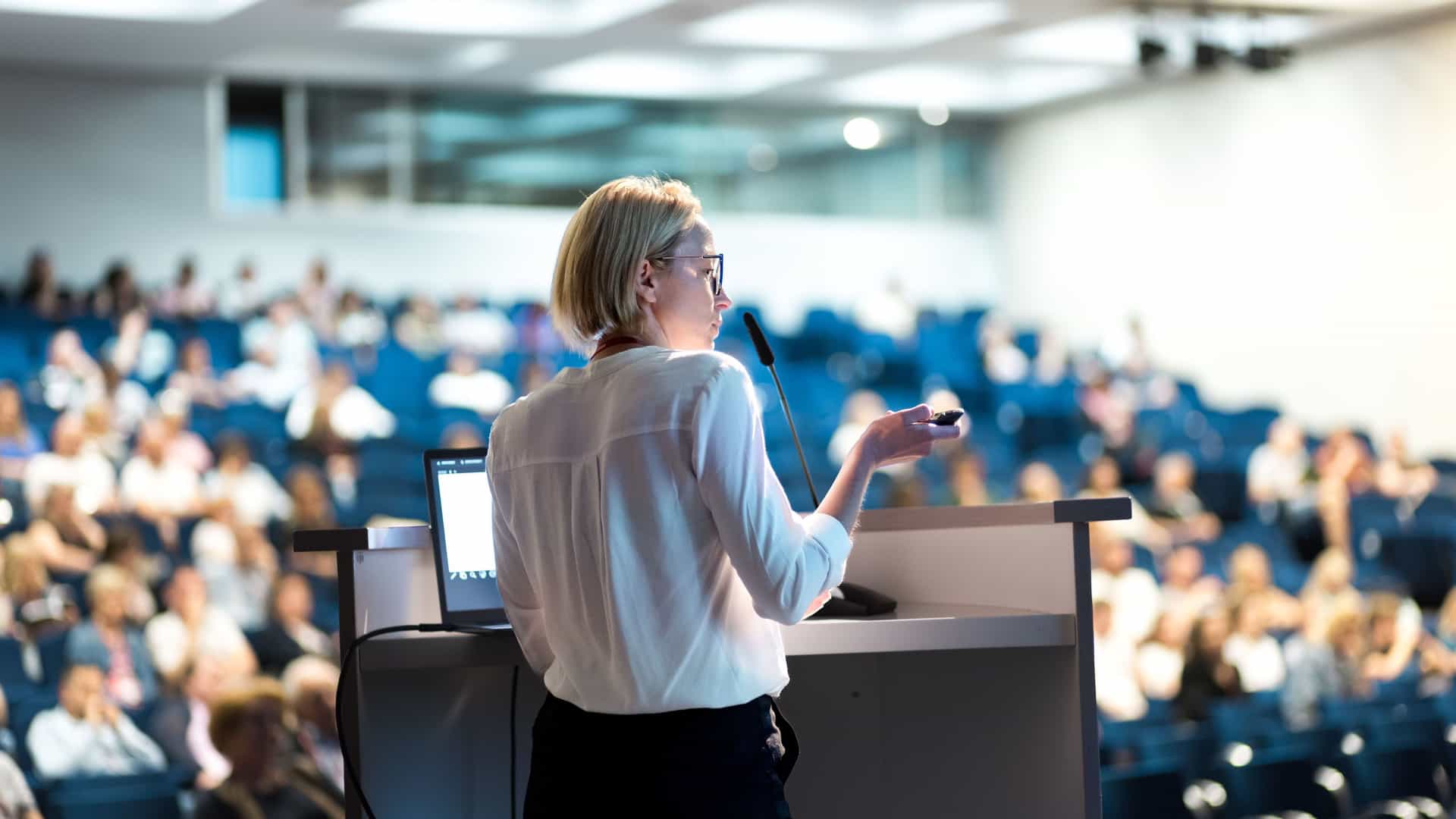 A rear view of a female lecturer with short blonde hair, glasses, and a white shirt standing at a podium. She holds a remote control in one hand and gestures with the other while speaking into a microphone. She is giving a presentation to a large, seated audience in a tiered auditorium. The audience is blurred in the background, focusing attention on the speaker. The scene represents an onboard enrichment program, a common feature of trans-Pacific cruises, which offers educational lectures to passengers.