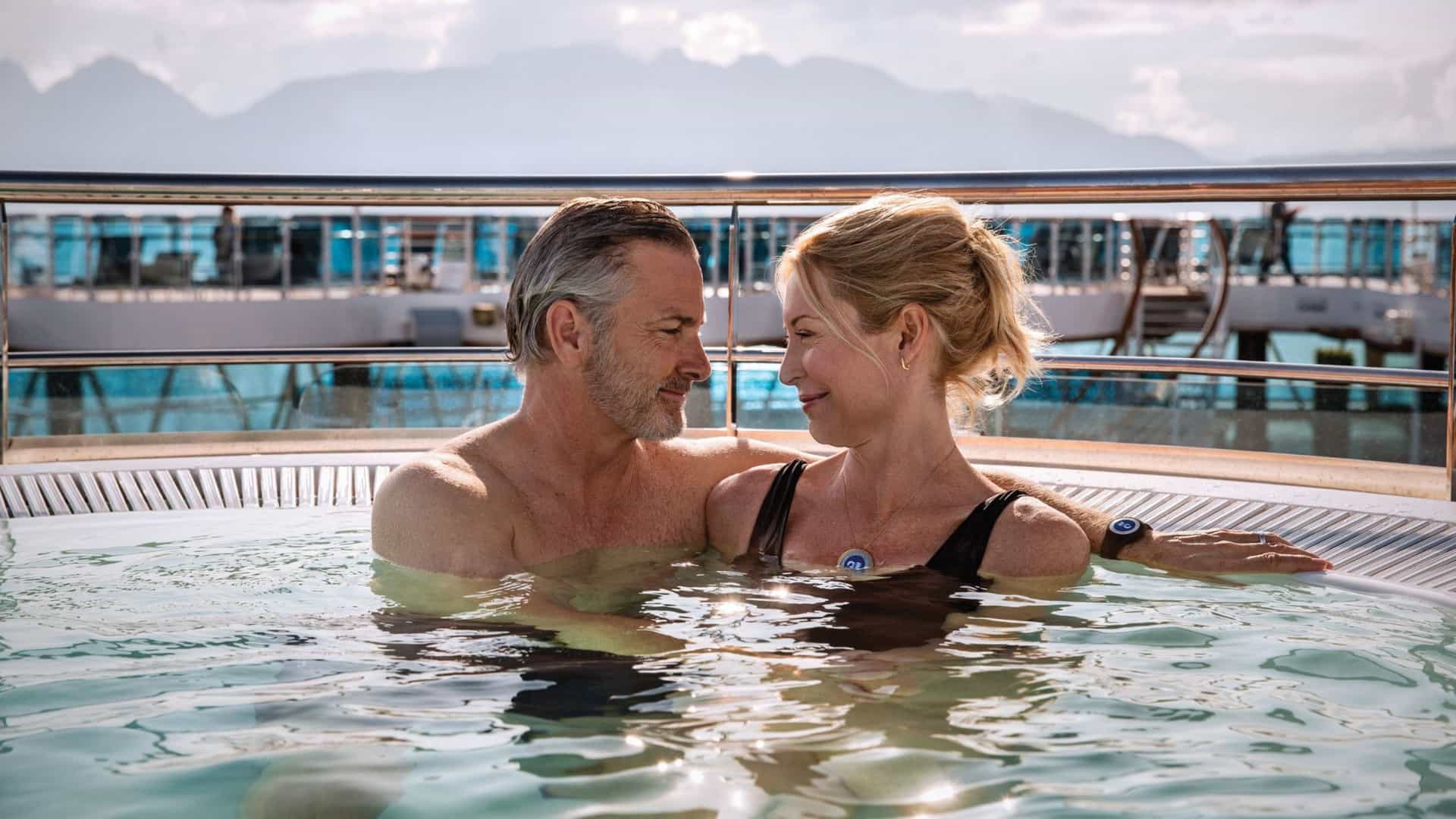A middle-aged couple is relaxing in a hot tub on the deck of a cruise ship. The man and woman are smiling and looking at each other, with the water bubbling around them. In the background, there is a balcony railing and mountains and a hazy sky are visible. The image captures a serene and romantic moment, showcasing the leisurely and intimate experience of a trans-Pacific cruise.