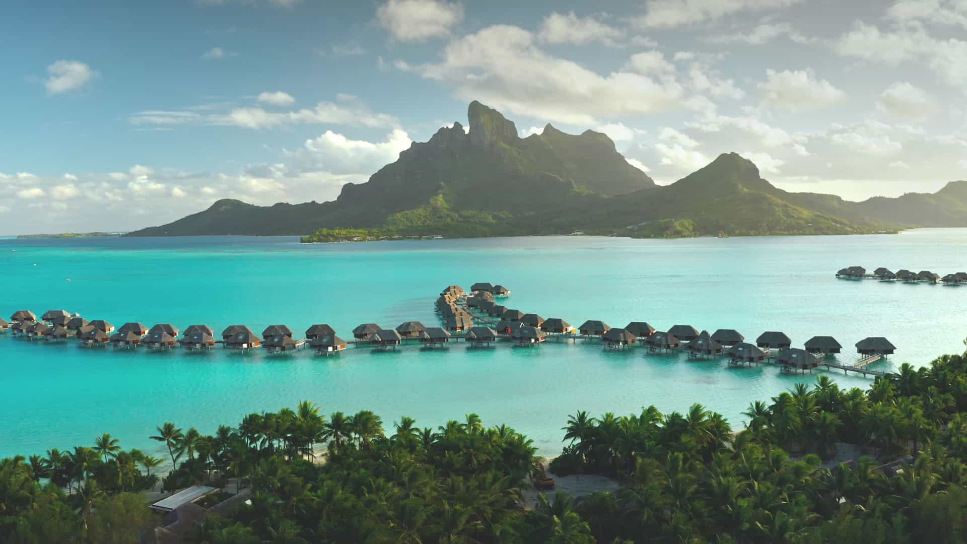 A panoramic view of luxurious overwater bungalows in a serene lagoon in Bora Bora, French Polynesia. The bungalows, with their thatched roofs, are built on stilts over the shallow, crystal-clear turquoise water. In the foreground, the tops of palm trees are visible. The central part of the image shows a long line of bungalows connected by a wooden pier. In the background, the majestic, lush green peaks of Mount Otemanu and Mount Pahia rise against a partly cloudy sky. The image captures the iconic resort and stunning natural beauty of this famous trans-Pacific destination.