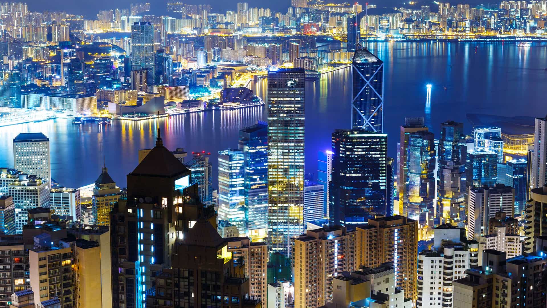 An iconic night view of the Hong Kong city skyline, a major destination on a trans-Pacific voyage. The city's modern skyscrapers are brilliantly lit against the dark night sky. The tall buildings of the financial district are visible, along with the glittering lights of the cityscape spreading across the hills. The dark water of the harbor reflects the city lights, creating a stunning visual display. The image captures the dynamic energy and modern urban landscape of this bustling Asian hub.