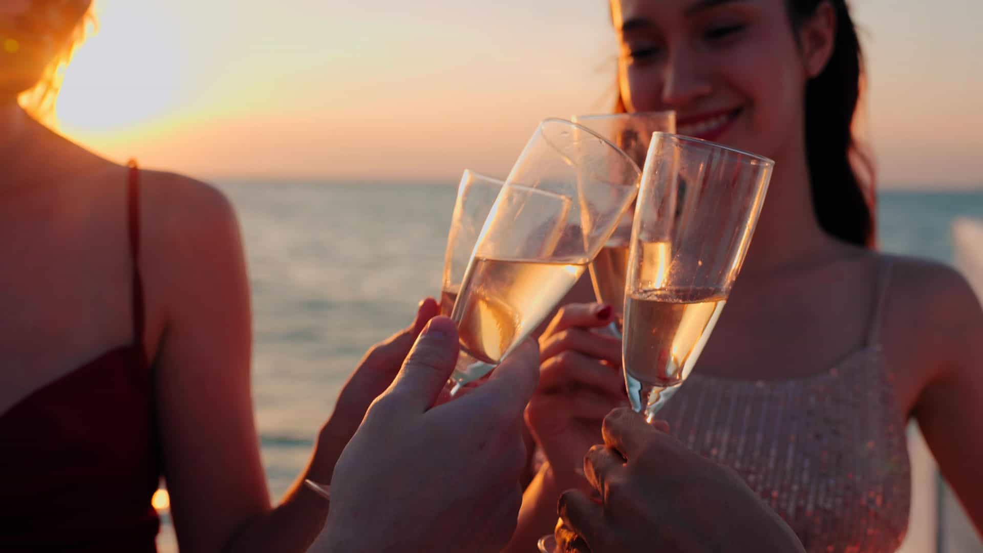 A group of people, appearing to be on a cruise ship, are celebrating with a champagne toast at sunset. Several hands holding champagne flutes are raised, clinking together in the center of the frame. The background shows the sparkling ocean and the vibrant orange and yellow hues of the setting sun, which casts a warm glow on the faces of the people. The image captures a celebratory and joyful moment during a journey across the Pacific Ocean.