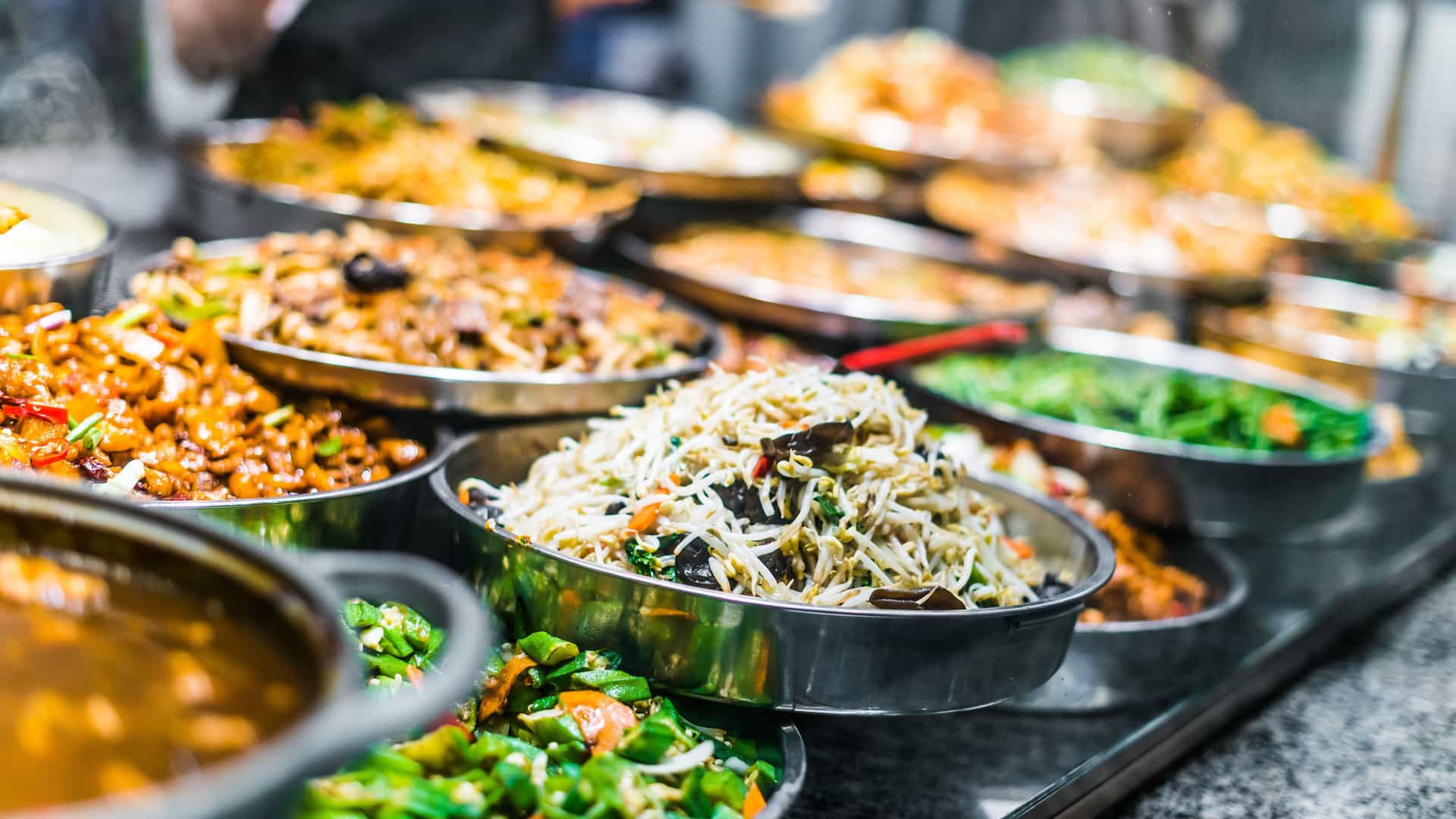 An abundant display of assorted Asian food dishes in a buffet-style restaurant. The food is arranged in large, circular metal serving bowls behind a glass counter. The dishes appear fresh and vibrant, including various noodle dishes with bean sprouts, stir-fried vegetables, and different types of curries and fried foods. The lighting highlights the textures and colors of the food, showcasing the variety and richness of Asian cuisine that might be available on a trans-Pacific cruise.