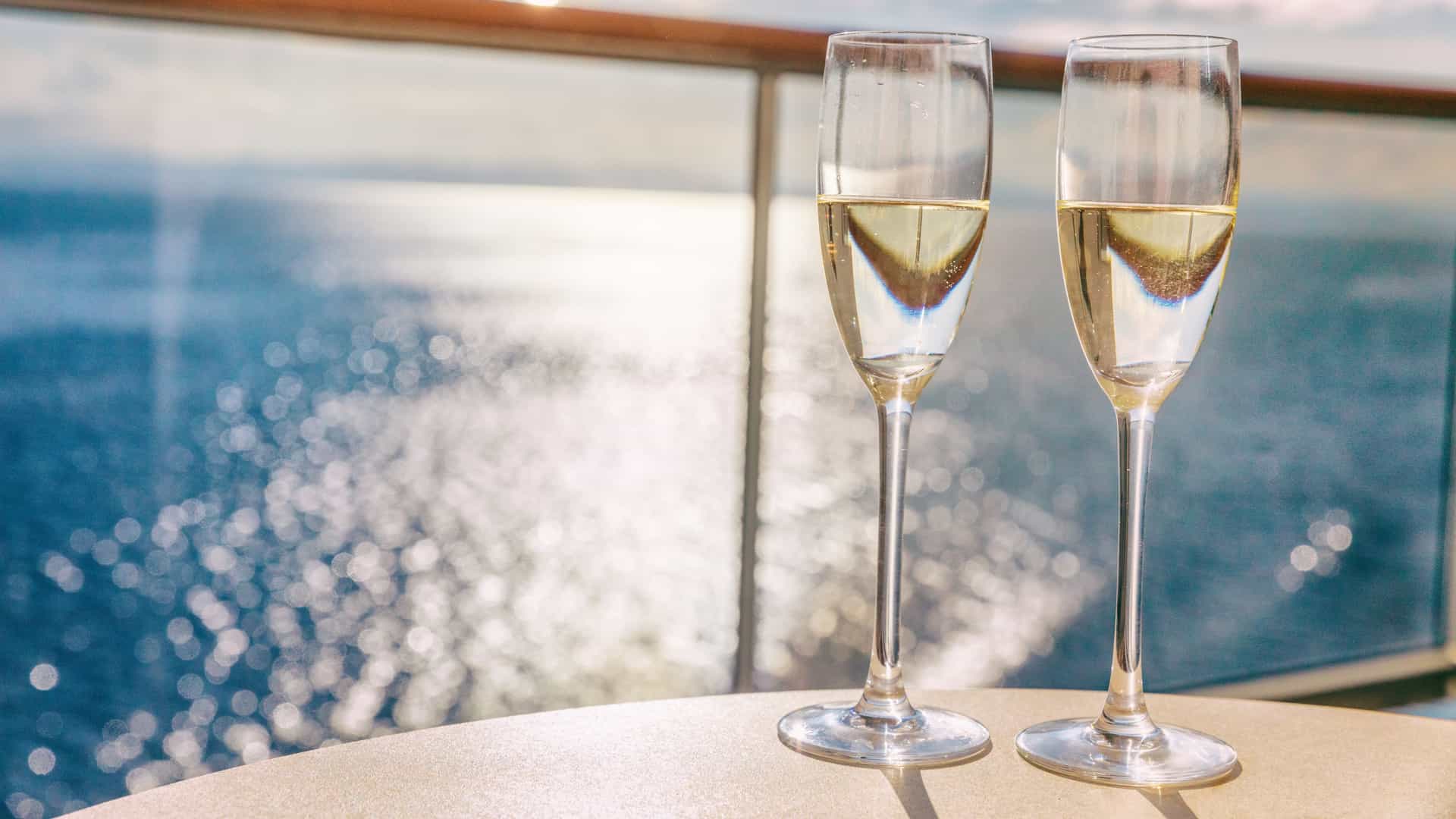 A close up shot of two champagne glasses on a private balcony aboard a cruise ship with the sun's reflection off the ocean surface in the background.