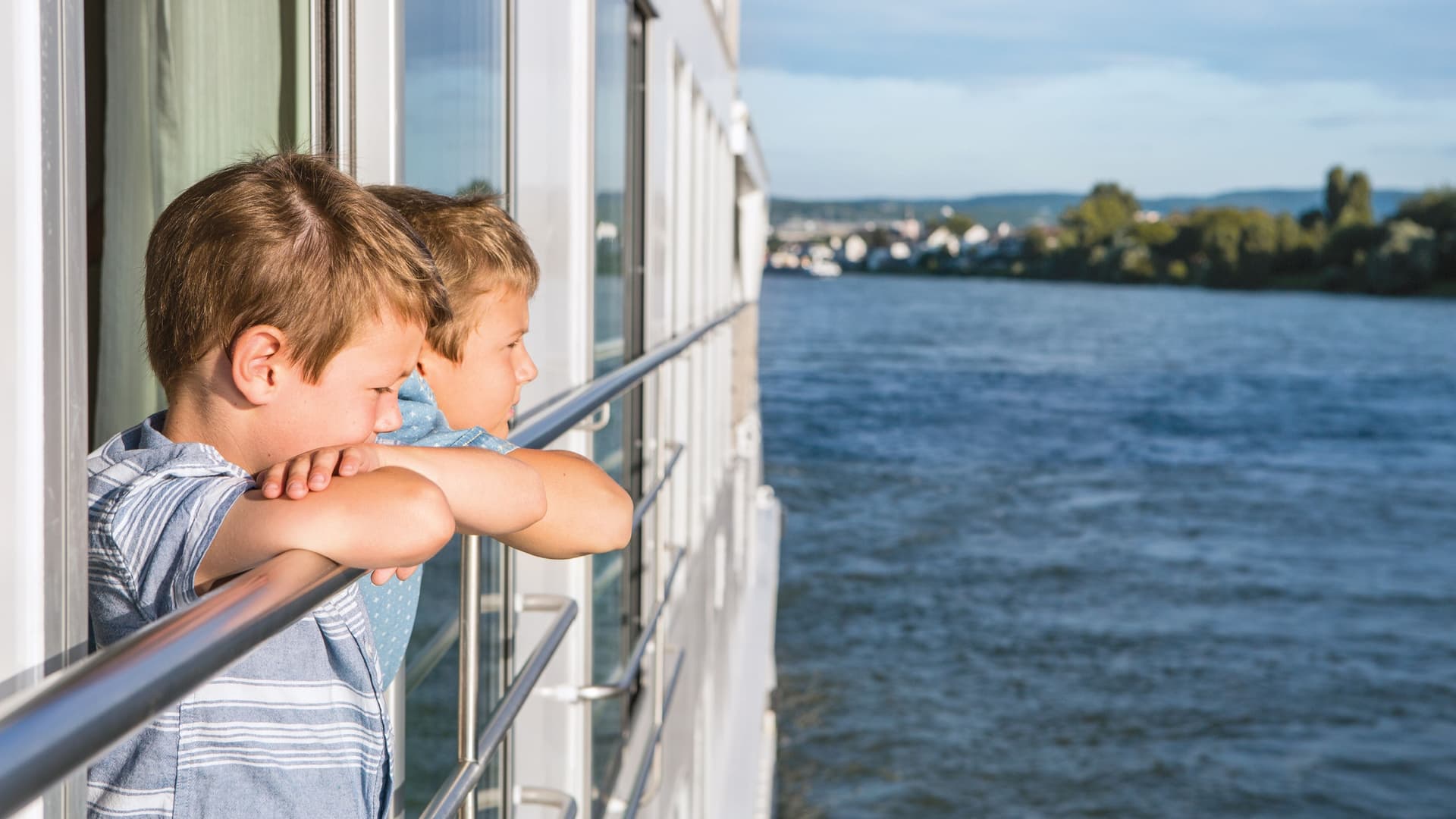 Two young boys lean on a Uniworld river cruise ship railing, looking out over a wide river toward a distant shoreline.