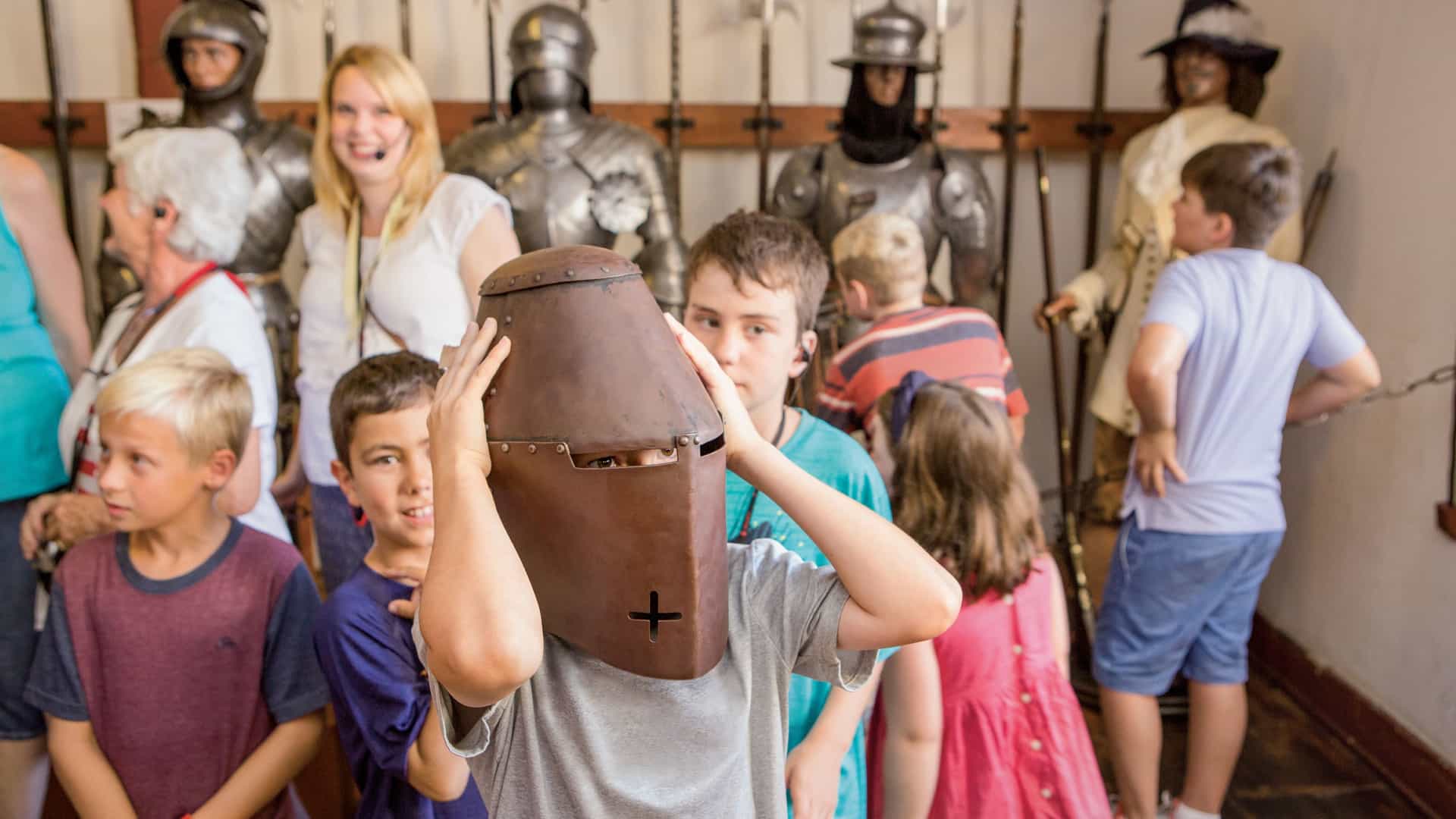 A young boy tries on a medieval knight's helmet during a group museum excursion surrounded by other children and suits of armor.