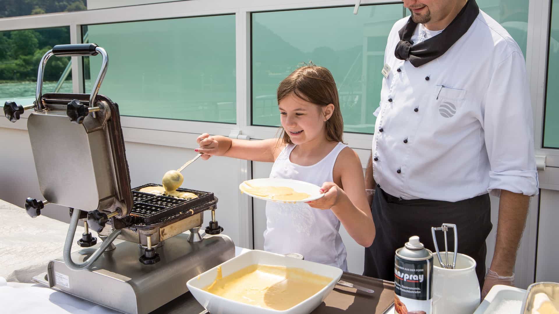 A young girl smiles as she pours batter onto an outdoor waffle iron on a cruise ship sundeck while a chef in uniform watches.