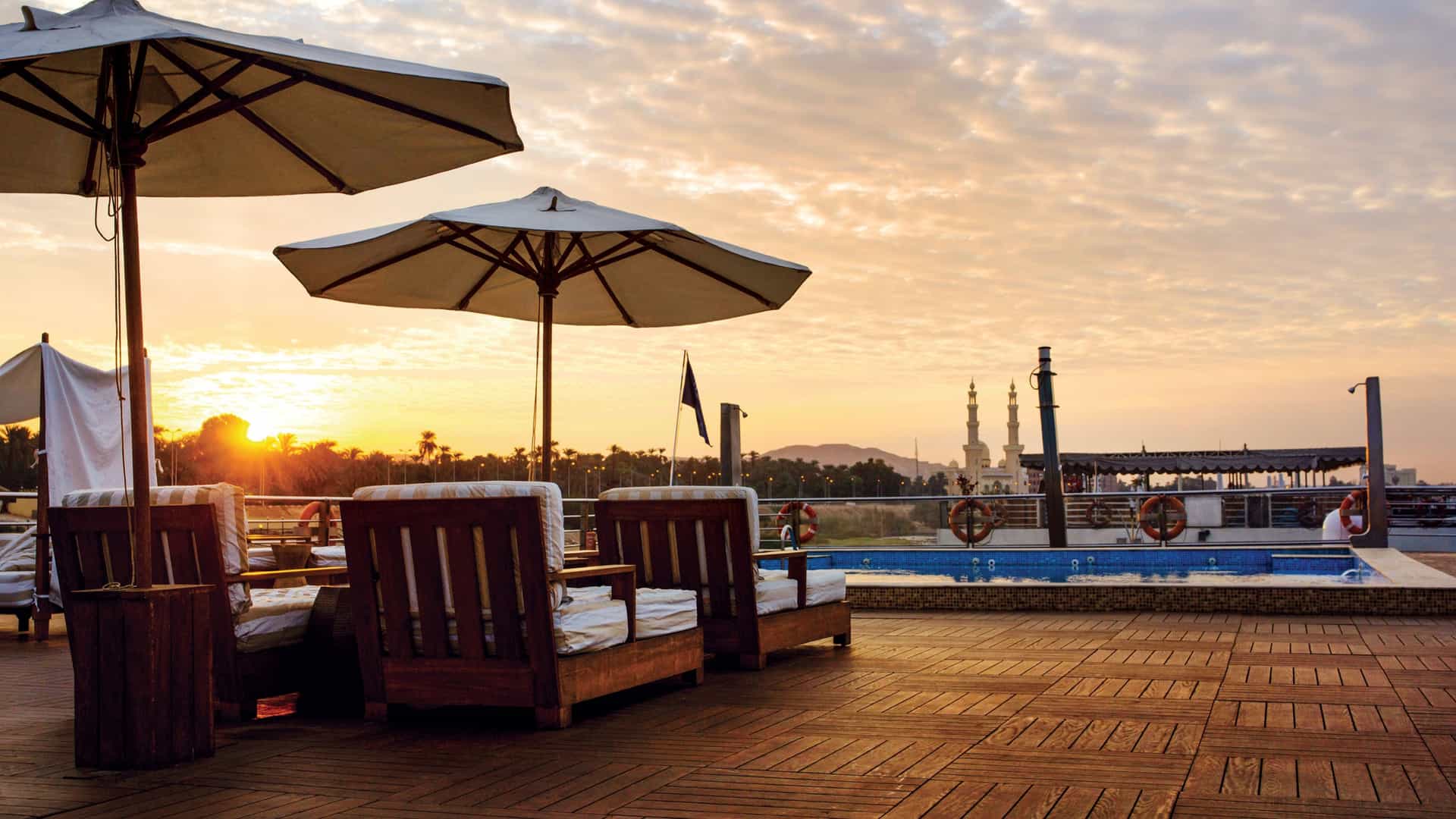 Wooden lounge chairs under umbrellas sit on a cruise ship's tiled sundeck next to a swimming pool, overlooking a river sunset and a distant mosque.