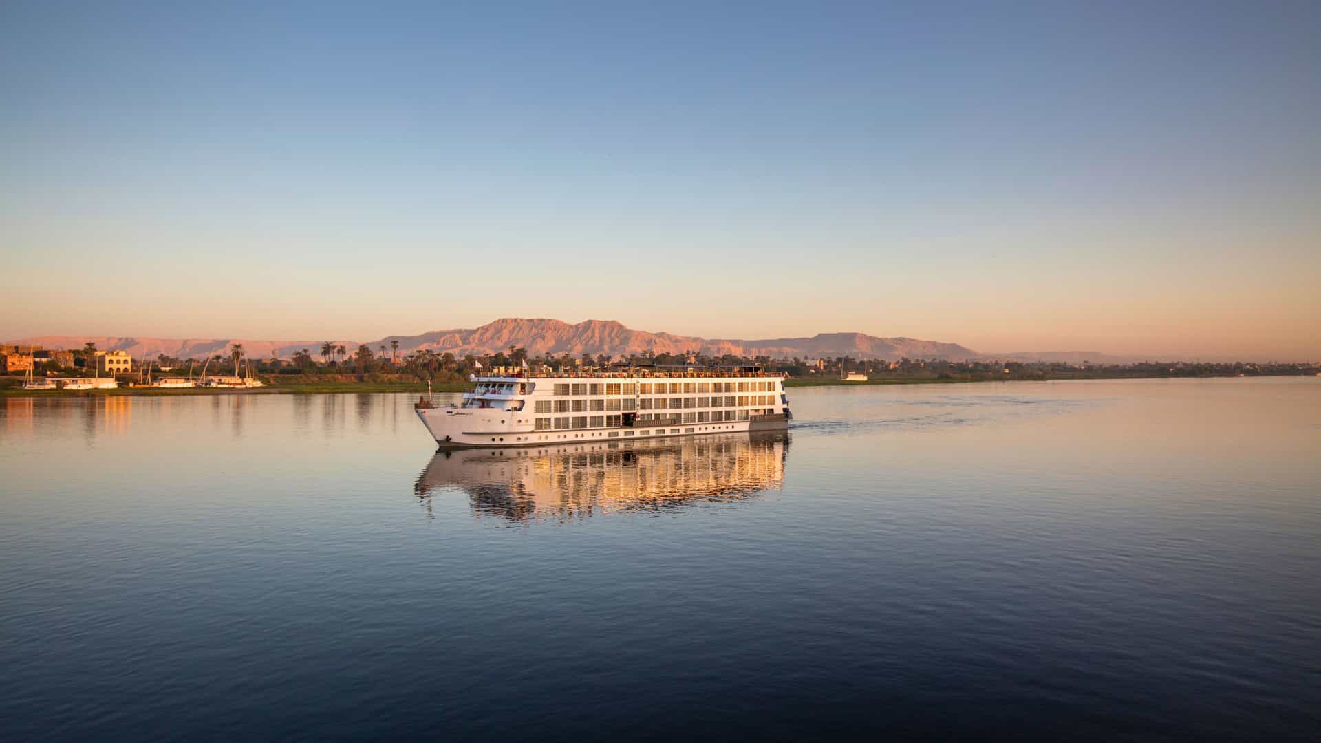A Uniworld cruise ship sailing on the calm waters of the Nile River in Egypt at sunset, with distant mountains and palm trees visible on the banks.