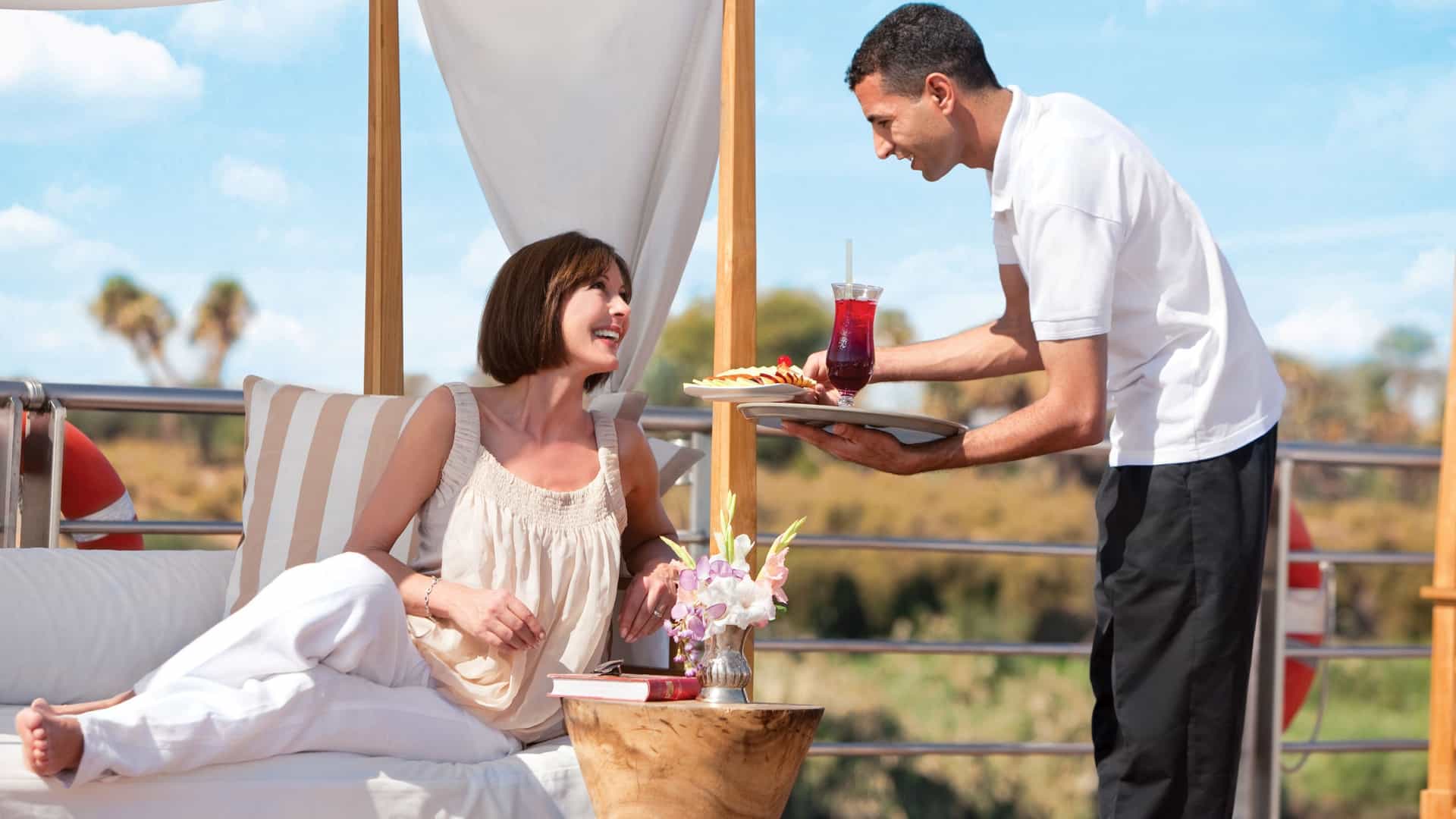 A woman being served sliced fruit and a cocktail on the rooftop sundeck of a Uniworld river ship.