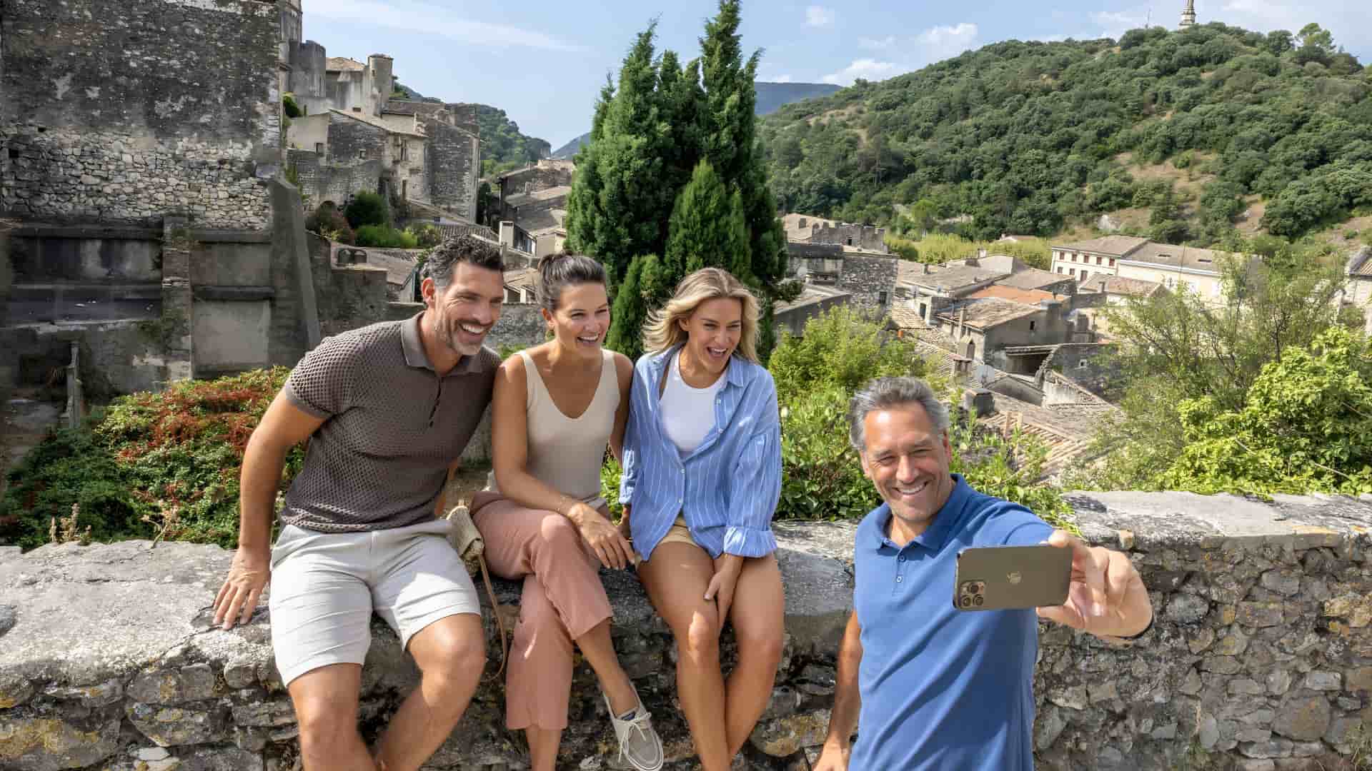 A group of four smiling travelers sitting on an ancient stone wall, posing for a selfie with a scenic backdrop of a historic European hillside village and lush greenery.