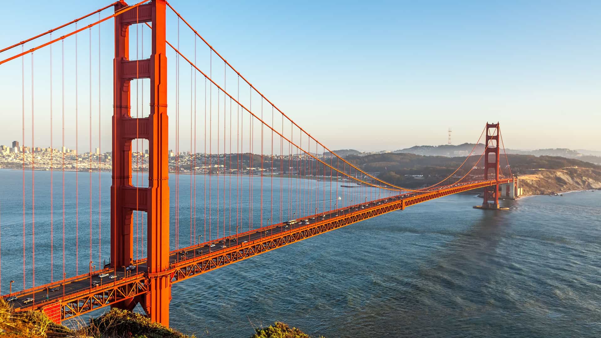 An iconic view of the Golden Gate Bridge in San Francisco, California, bathed in the warm light of late afternoon. The bridge's signature international orange towers and suspension cables span across the dark blue waters of the bay. On the left, the city skyline of San Francisco is visible in the distance, while on the right, the rolling green hills of Marin County lead to the bridge's northern tower. The image captures the famous landmark along the U.S. Pacific Coast.