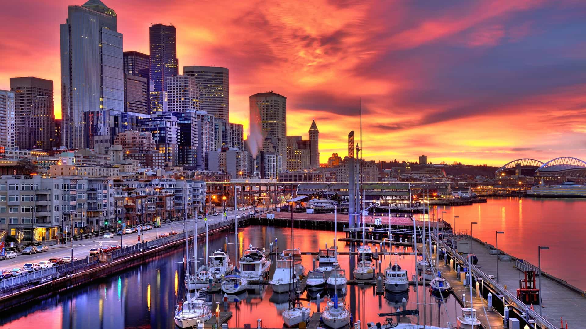 A dramatic sunset over the city of Seattle, Washington. The sky is a fiery mix of orange, red, and purple clouds, casting a vibrant glow over the cityscape. In the foreground, a marina is filled with sailboats and yachts, their masts reflecting in the calm harbor water. The downtown skyline, with its prominent skyscrapers, rises behind the marina. The image captures the striking urban landscape of a major city on the U.S. Pacific Coast.