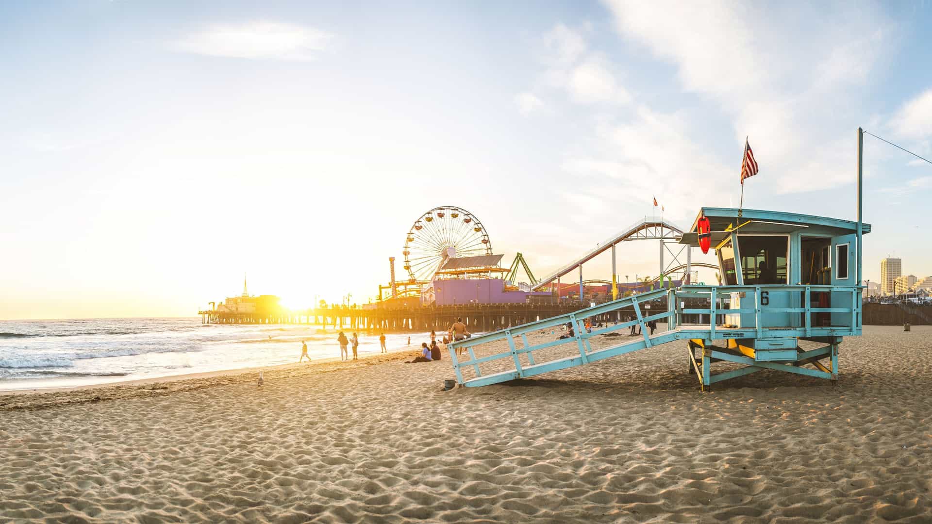 A wide-angle, panoramic view of the beach at Santa Monica, California, at sunset. The iconic light-blue lifeguard tower stands in the foreground on the sandy beach. In the distance, the Santa Monica Pier is silhouetted against the setting sun, with its Ferris wheel and roller coaster visible. The calm waves of the Pacific Ocean lap at the shore, and a few people can be seen walking along the beach. The image captures the relaxed, coastal atmosphere of a famous spot on the U.S. Pacific Coast.