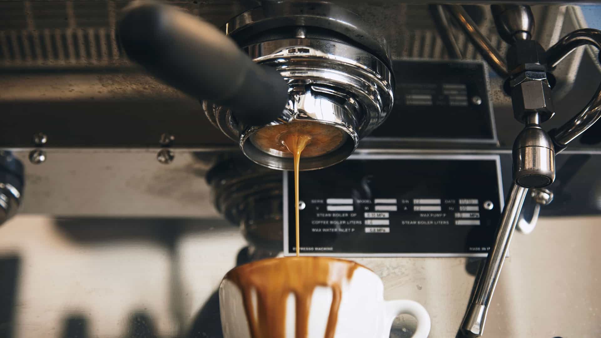 A close-up of a professional espresso machine as it brews a shot of coffee. A stream of rich, dark espresso pours from the portafilter into a white ceramic cup. The machine's polished chrome and stainless steel components are visible, creating a sleek, industrial aesthetic. The image captures the meticulous process of coffee making, a significant part of the artisan food and drink culture found in many cities along the U.S. Pacific Coast.