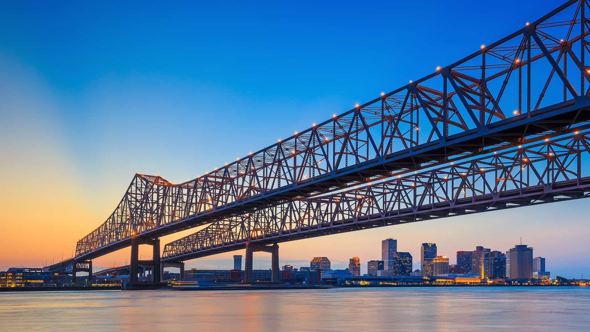New Orleans skyline reflected in calm water at sunset, showcasing tall buildings and vibrant colors.