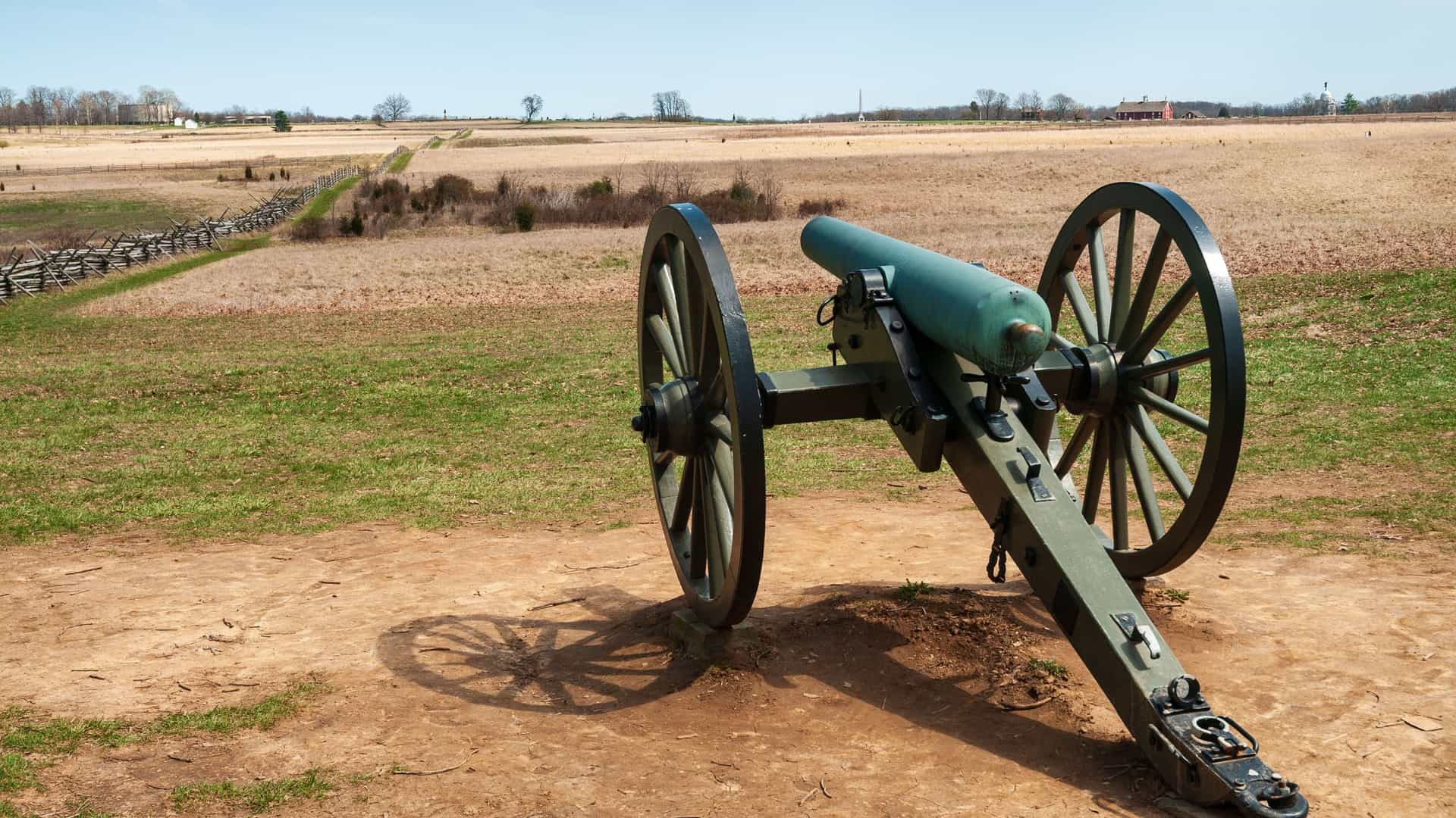A historic civil war cannon sits in a vast, open field at Gettysburg National Military Park. The cannon, painted an olive green, is positioned on a slight hill, with its wooden wheels prominent in the foreground. In the background, a long wooden fence runs diagonally across the field, leading toward a cluster of trees in the distance. The landscape is a mix of dry, golden-brown grass and patches of green, with a clear blue sky above. The image depicts a significant historical site along a U.S. river route.