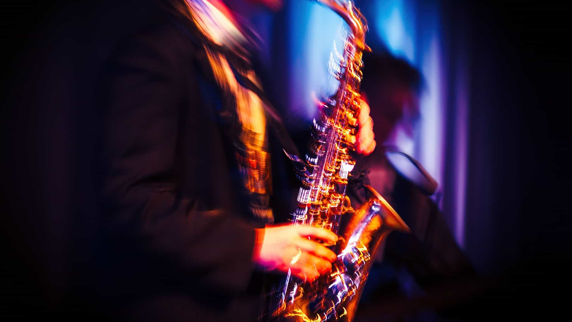 A motion-blurred photograph of a jazz musician playing a saxophone in a dimly lit club. The musician's face is a blur of motion, but their hands are visible on the keys of the golden brass instrument, which glows with reflected light. The background is dark, with a vertical blue light source on the right, suggesting a stage or a spotlight. The image captures the energetic and improvisational spirit of live jazz, a genre deeply associated with cities like New Orleans on the Mississippi River.