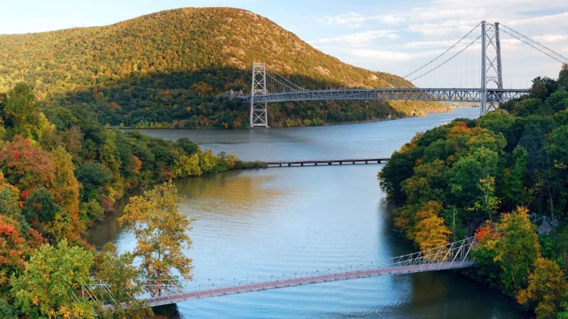 An autumn view of the Hudson River with the Bear Mountain Bridge. The river flows through a valley surrounded by hills covered in trees showing a mix of green, yellow, and orange foliage. The large suspension bridge spans the river connecting the two forested shores. A smaller, lower bridge is also visible. The image captures the scenic beauty of the Hudson River Valley in New York, a major U.S. river region.