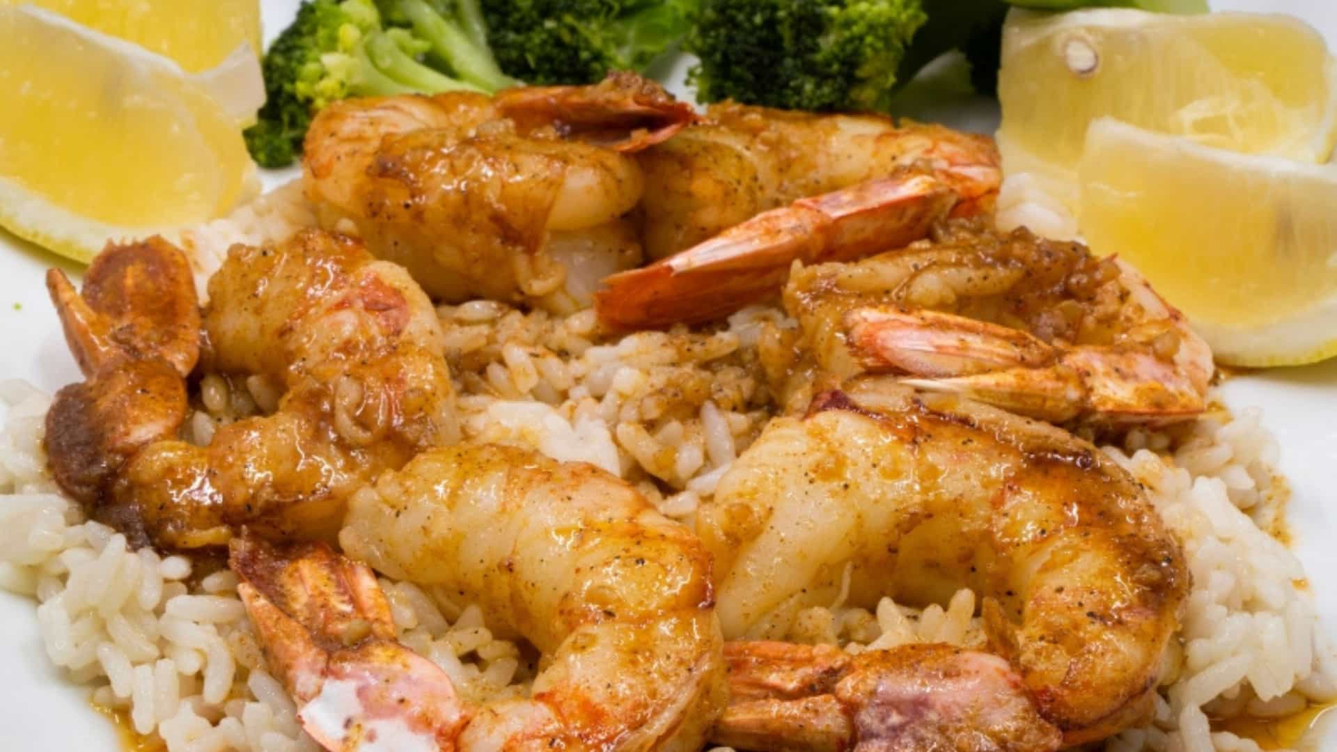 A plate of traditional Louisiana Creole or Cajun shrimp and rice. The dish features several large, succulent shrimp, seasoned and cooked in a flavorful sauce, served on a bed of fluffy white rice. A side of steamed green broccoli florets and several bright yellow lemon wedges garnish the plate. The image captures a classic and delicious meal from the cuisine of the Mississippi River region.