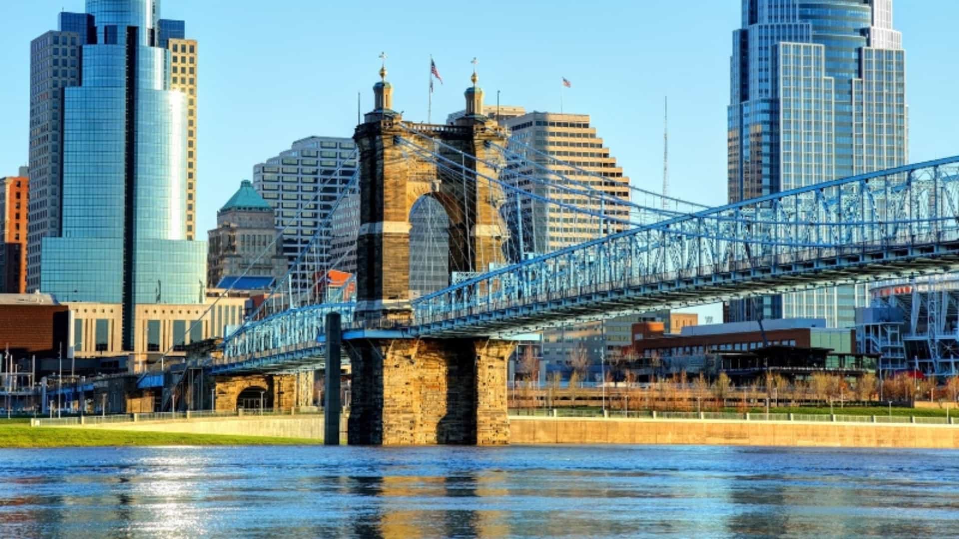 The John A. Roebling Suspension Bridge spans the Ohio River, connecting Cincinnati, Ohio, and Covington, Kentucky. The bridge's intricate steel suspension system and large stone towers are the focal point of the image. The Cincinnati skyline, featuring modern skyscrapers and office buildings, is visible in the background against a clear, blue sky. The calm surface of the Ohio River reflects the sunlight, creating a serene urban landscape. The image showcases a key feature of a major U.S. river city.
