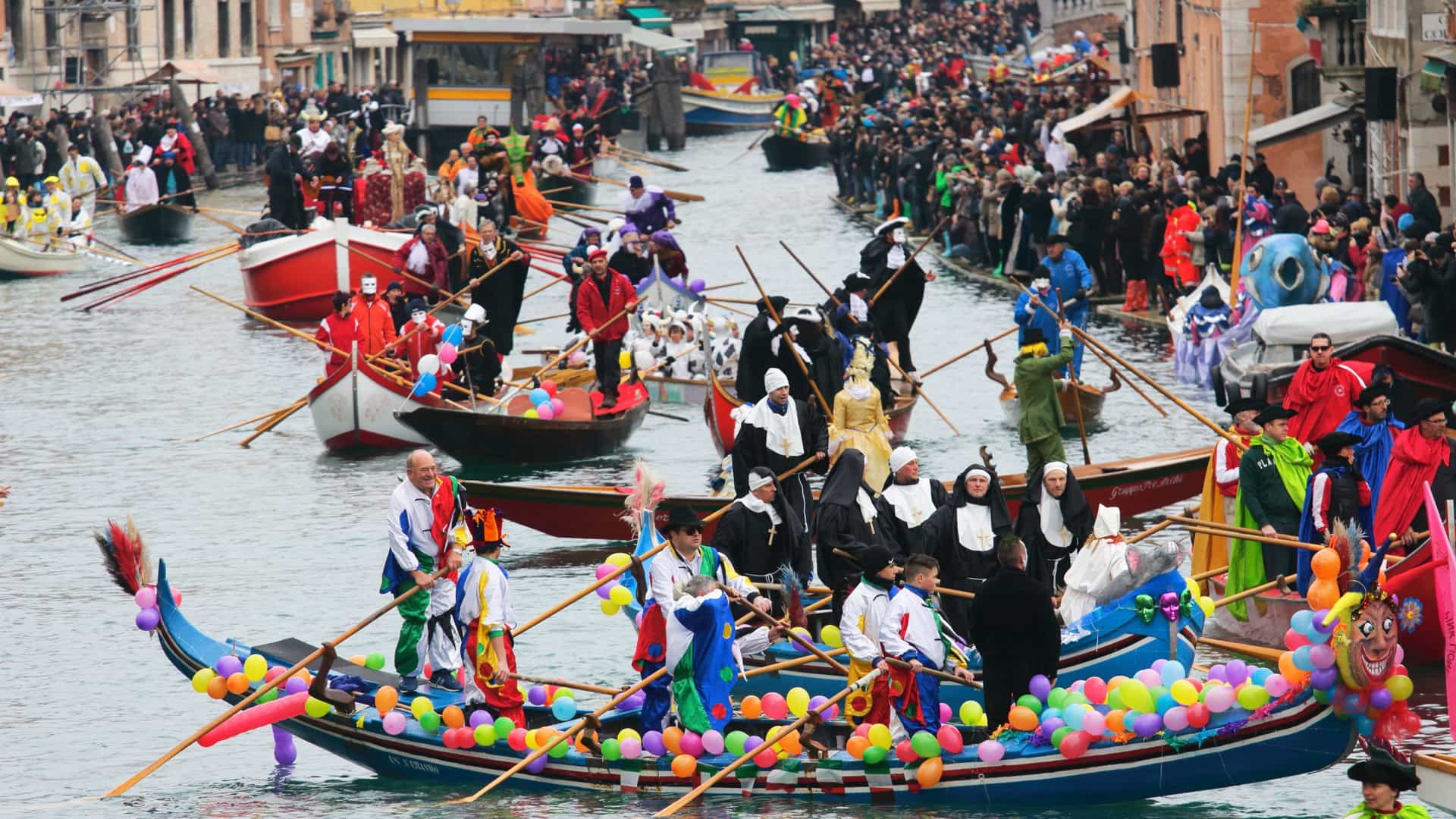 A festive scene on a Venetian canal shows a colorful boat parade during a celebration, the Venice Carnival.