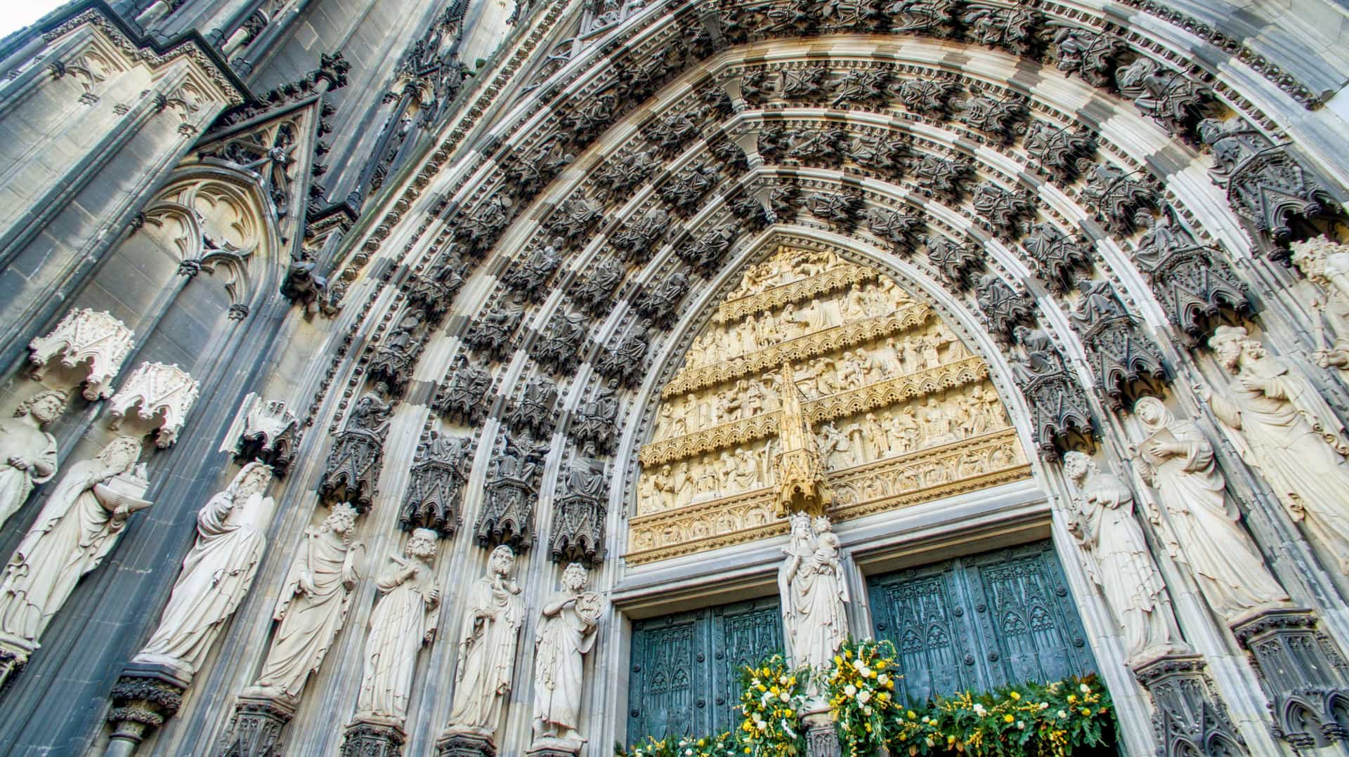 A detailed architectural close-up of the Gothic stone entrance arch and facade of Cologne Cathedral, decorated with carved statues and golden relief work.