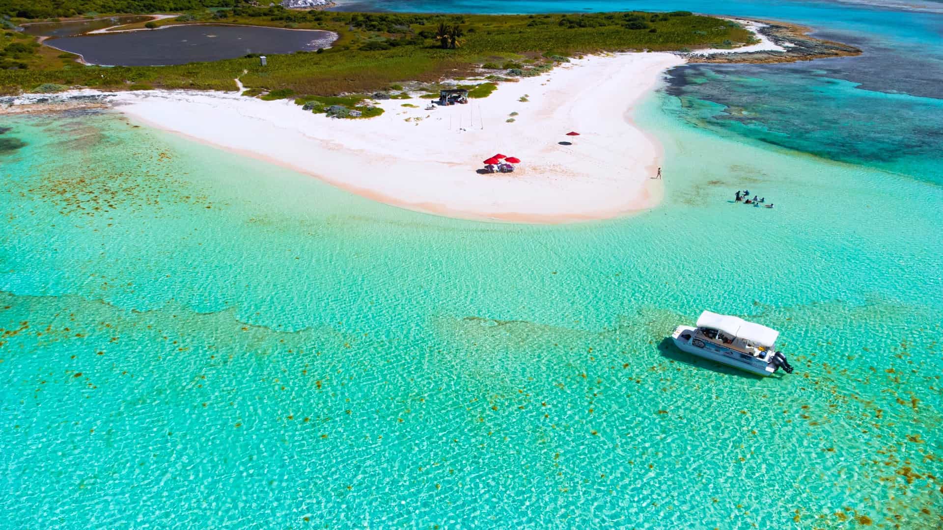 Aerial view of San Salvador Island in The Bahamas with white beaches, turquoise waters, a boat near shore, and red umbrellas—perfect for Viking Expedition Cruise travelers.
