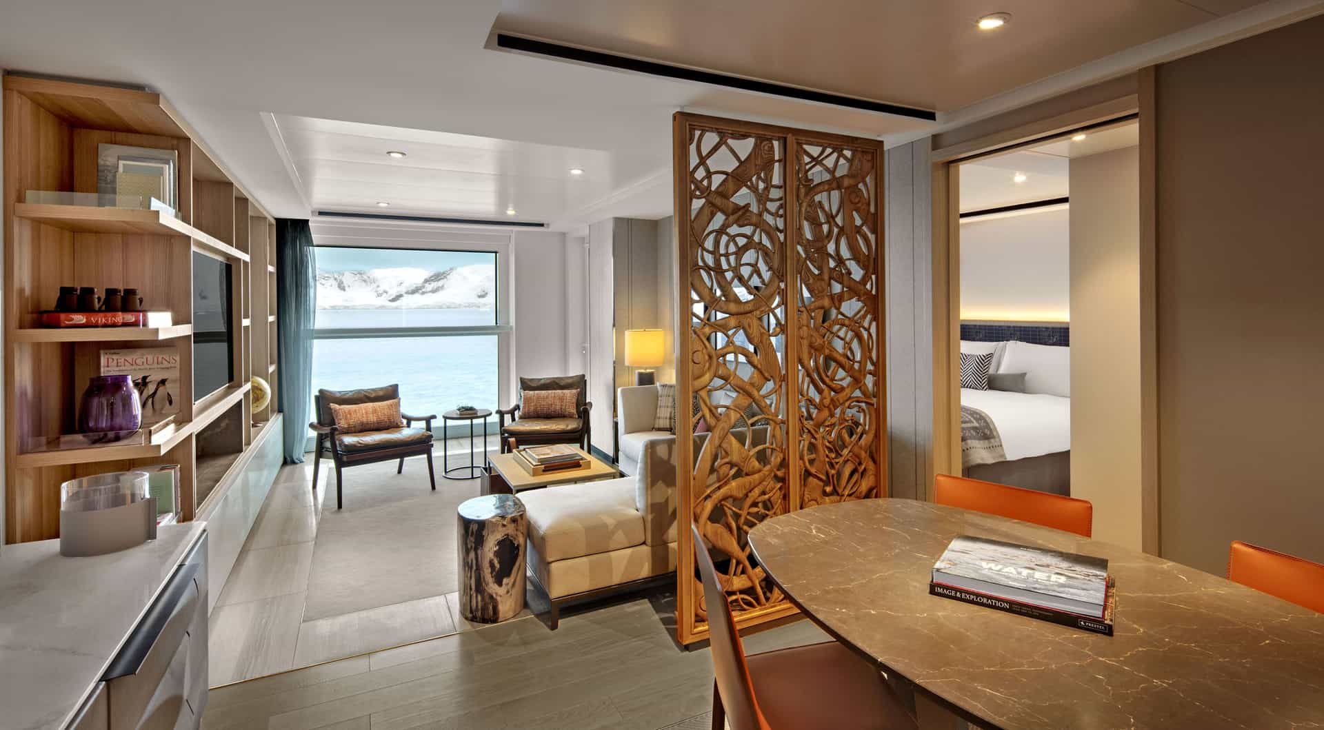A spacious shipboard suite features a dining area, a living room separated by an intricately carved wooden screen, and a large window facing snowy mountains.