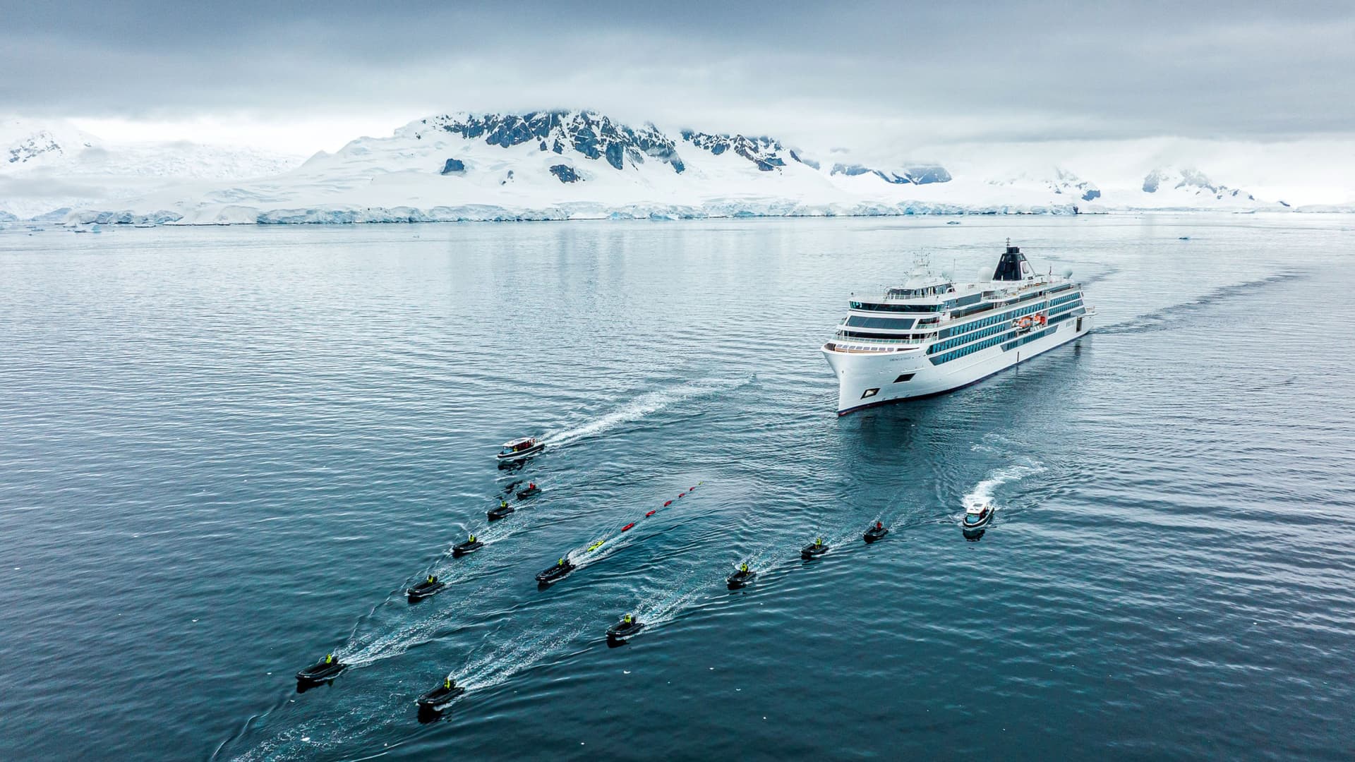 A white Viking expedition cruise ship sails through calm, icy polar waters, accompanied by a fleet of small Zodiac boats leaving wakes in front of snow-capped mountains.