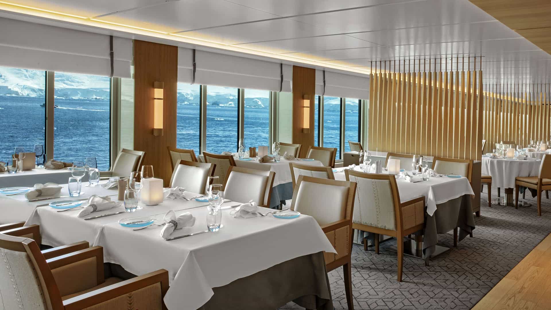 An elegant shipboard dining room with crisp white-clothed tables, Scandinavian wood accents, and large windows revealing views of icy ocean waters.