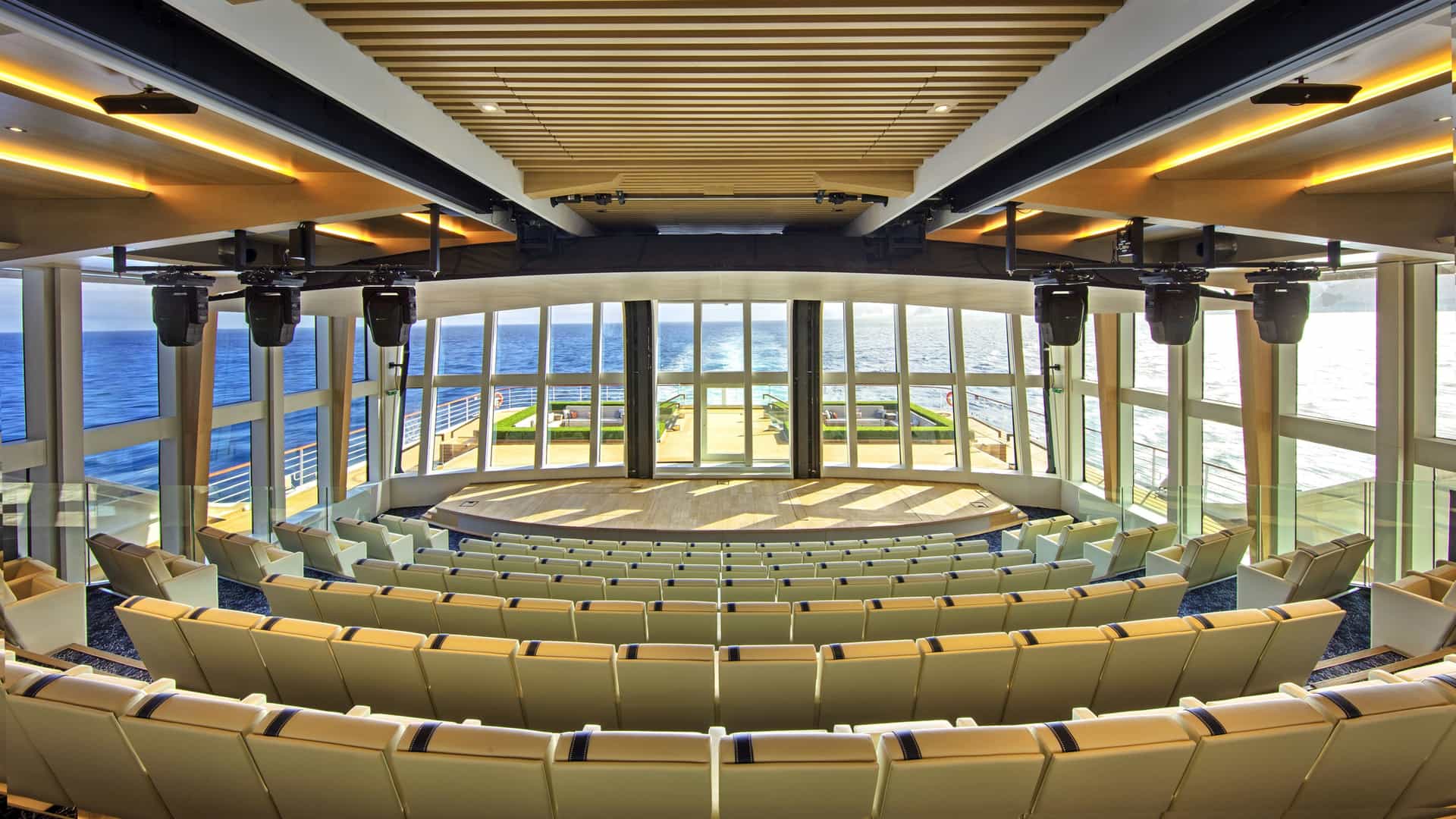 A modern, tiered auditorium onboard a ship features light leather seating and floor-to-ceiling glass windows offering panoramic ocean views.